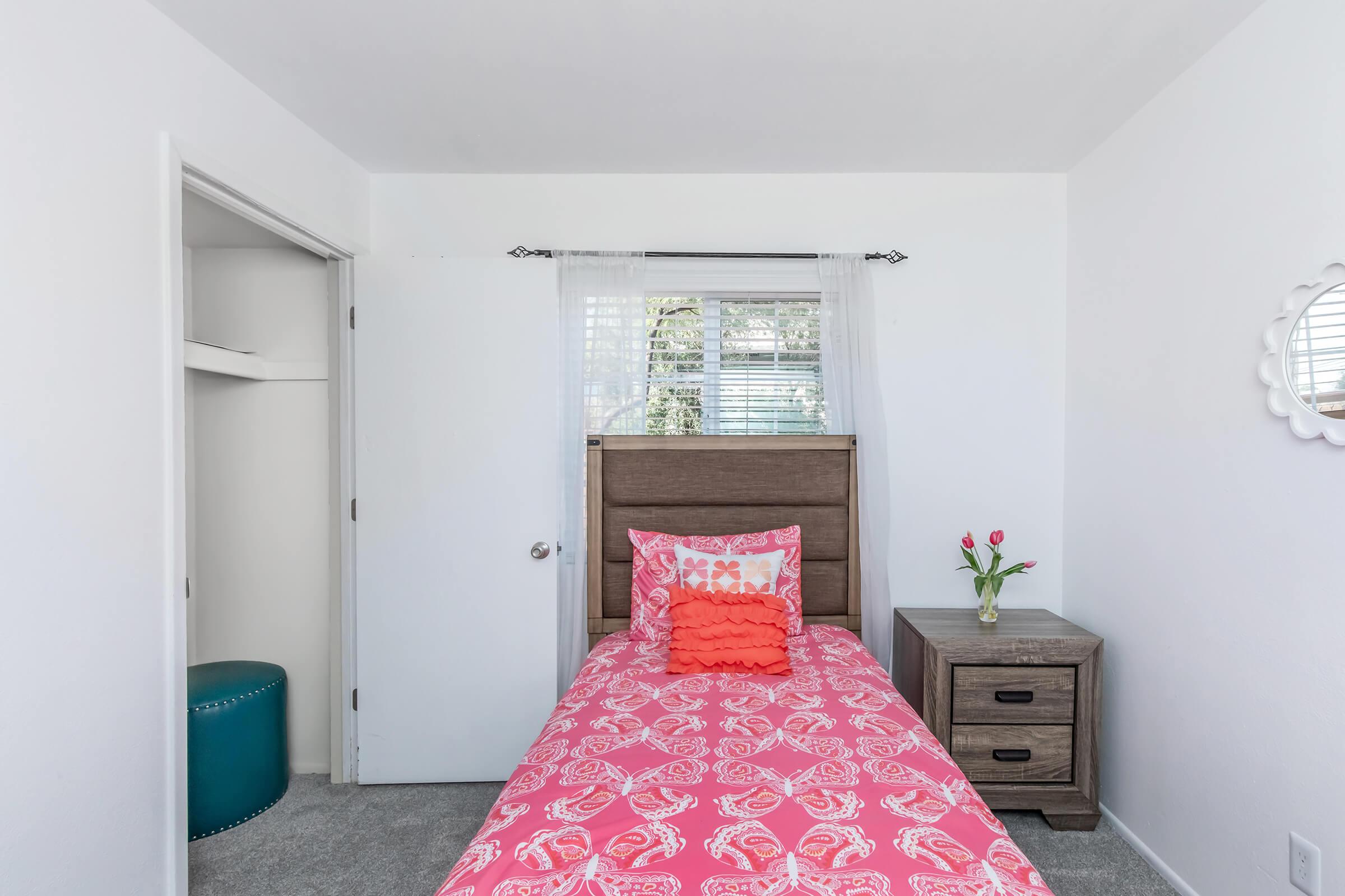 A bright bedroom featuring a single bed with a pink floral bedspread, a wooden headboard, and decorative pillows. There is a small nightstand with a lamp, a turquoise stool nearby, and a closet with an open door. Natural light streams in from a window covered with sheer curtains.