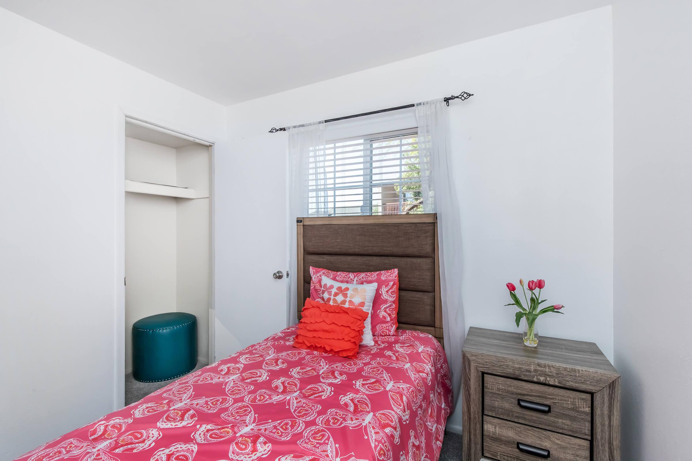 A cozy bedroom featuring a single bed with a bright pink and patterned comforter, a decorative pillow, and a small bedside table. There is a window with sheer curtains allowing natural light, and a vase of tulips on the table. A closet is partially visible in the background.
