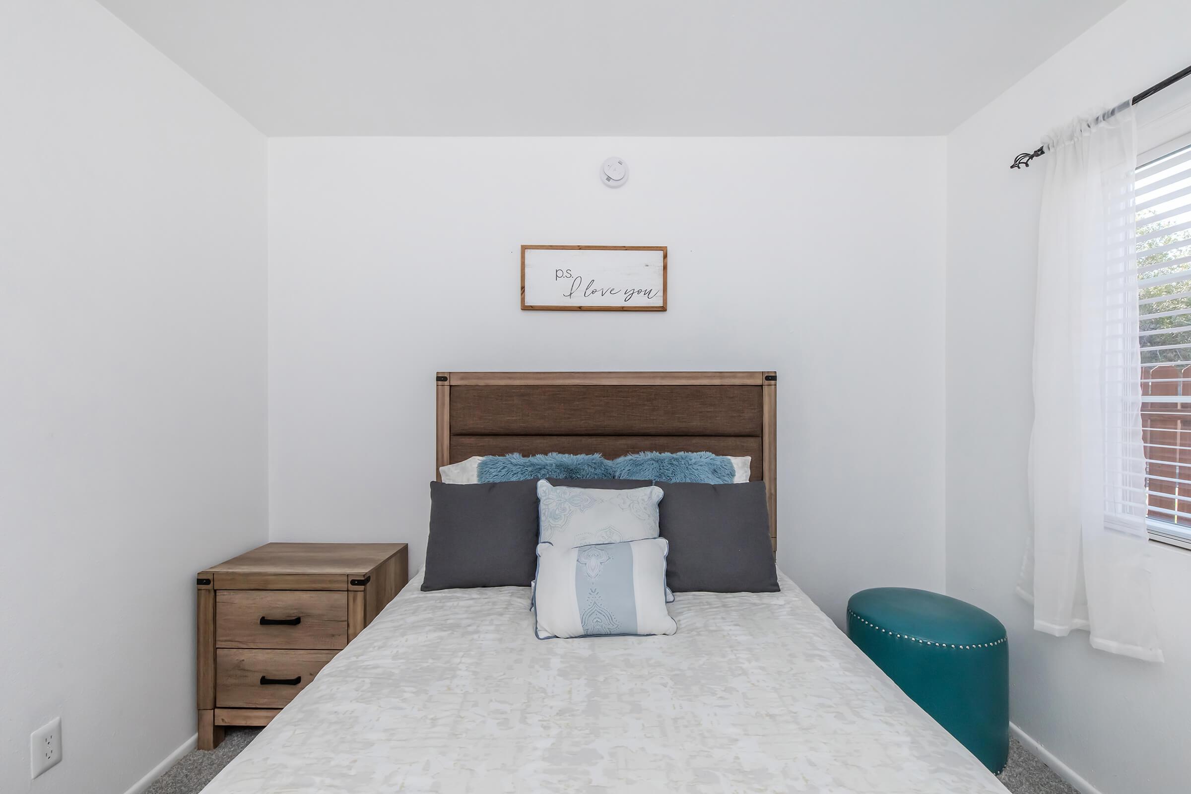 A cozy bedroom featuring a light-colored bed with decorative pillows, a wooden headboard, and a bedside table. A round teal ottoman sits beside the bed. The room has white walls and a window with sheer curtains, allowing natural light to brighten the space. A decorative sign on the wall adds a personal touch.