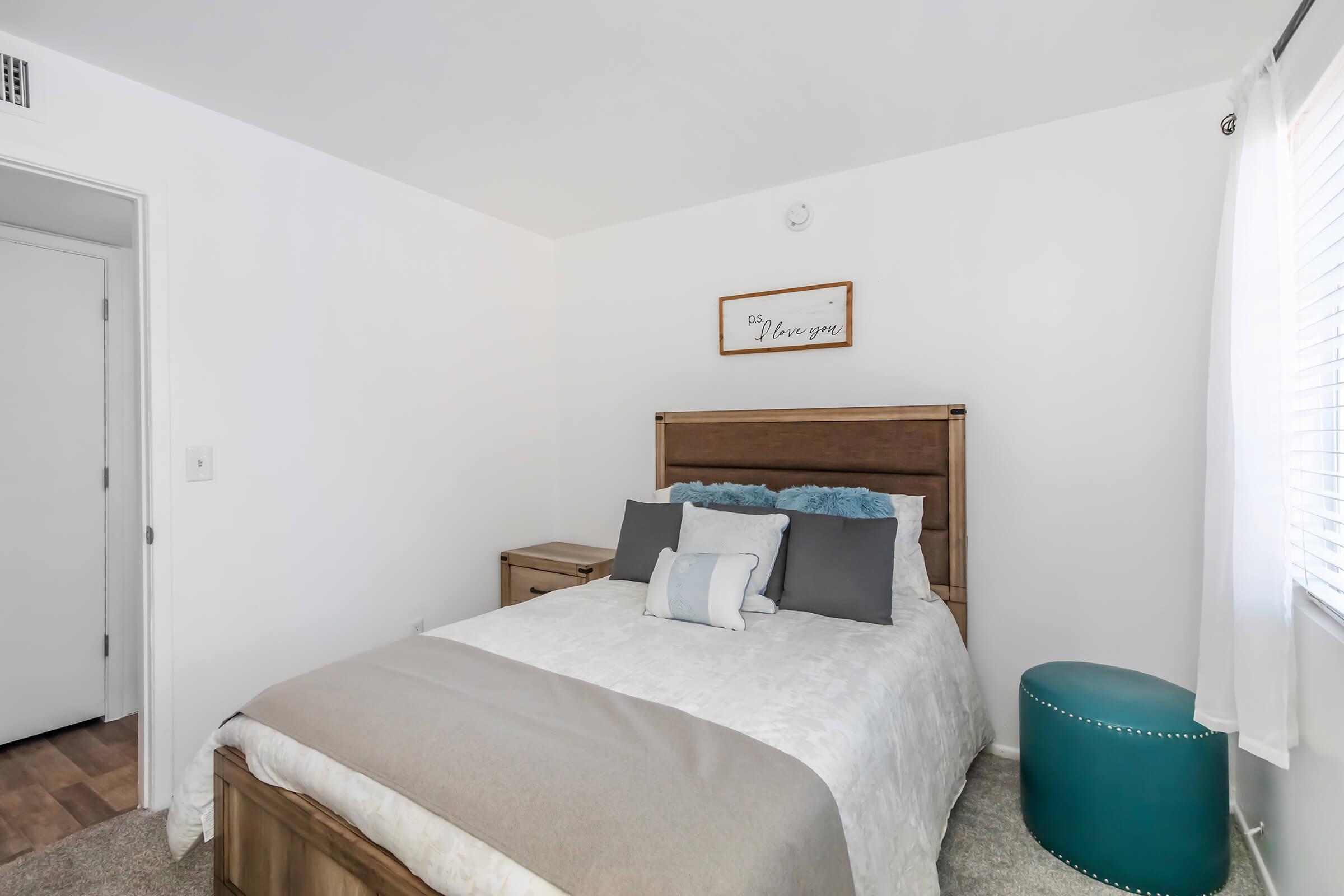 A cozy bedroom featuring a wooden bed frame with decorative pillows, a light gray bedspread, and a round teal nightstand. The room has white walls and a window with curtains, creating a bright and inviting atmosphere. A sign on the wall reads "The Message."