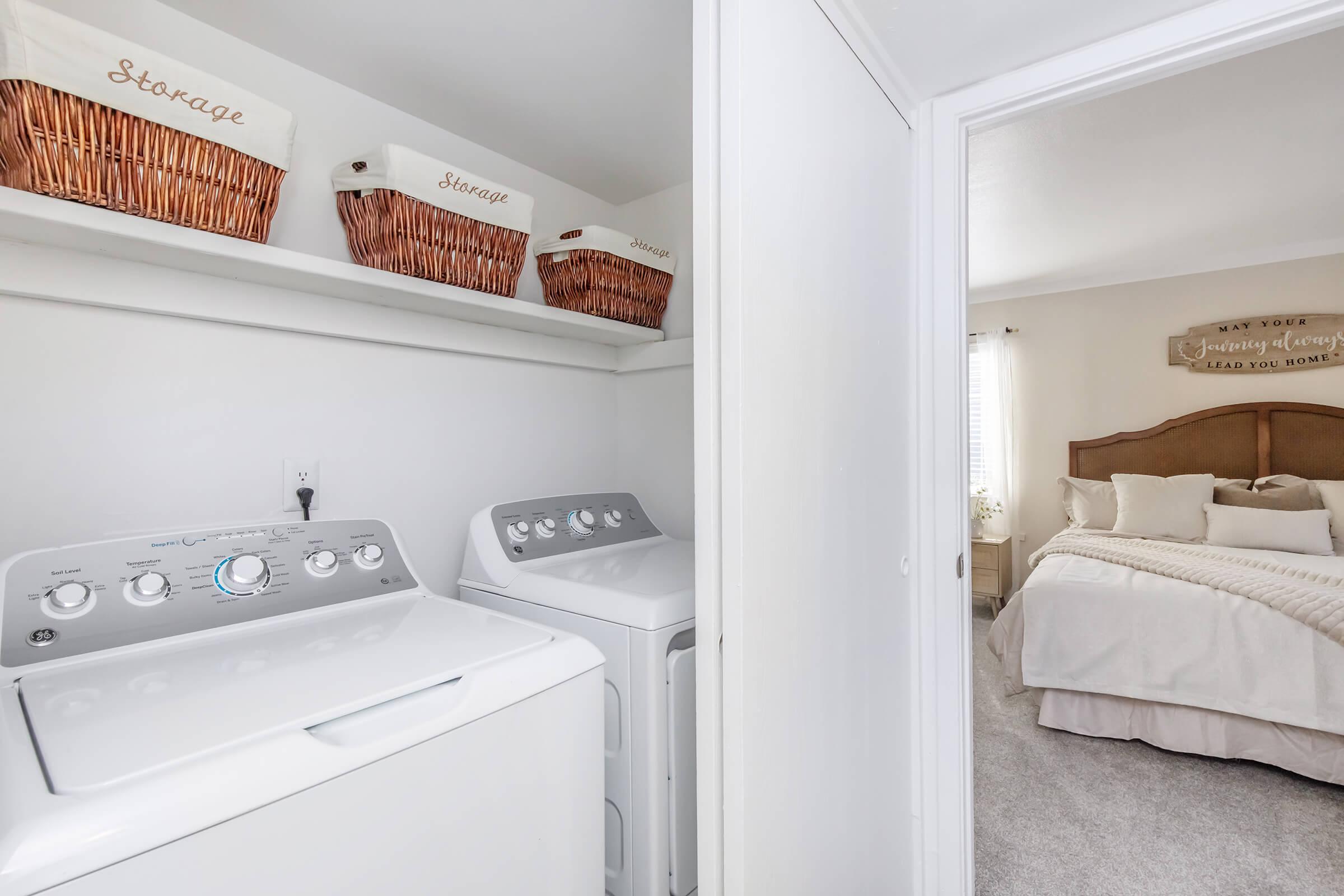 A laundry area featuring stacked washing machines and neatly organized wicker storage baskets on a shelf. The space connects to a bright bedroom with a cozy bed adorned with light-colored bedding and decorative pillows, enhancing the inviting atmosphere of the home.