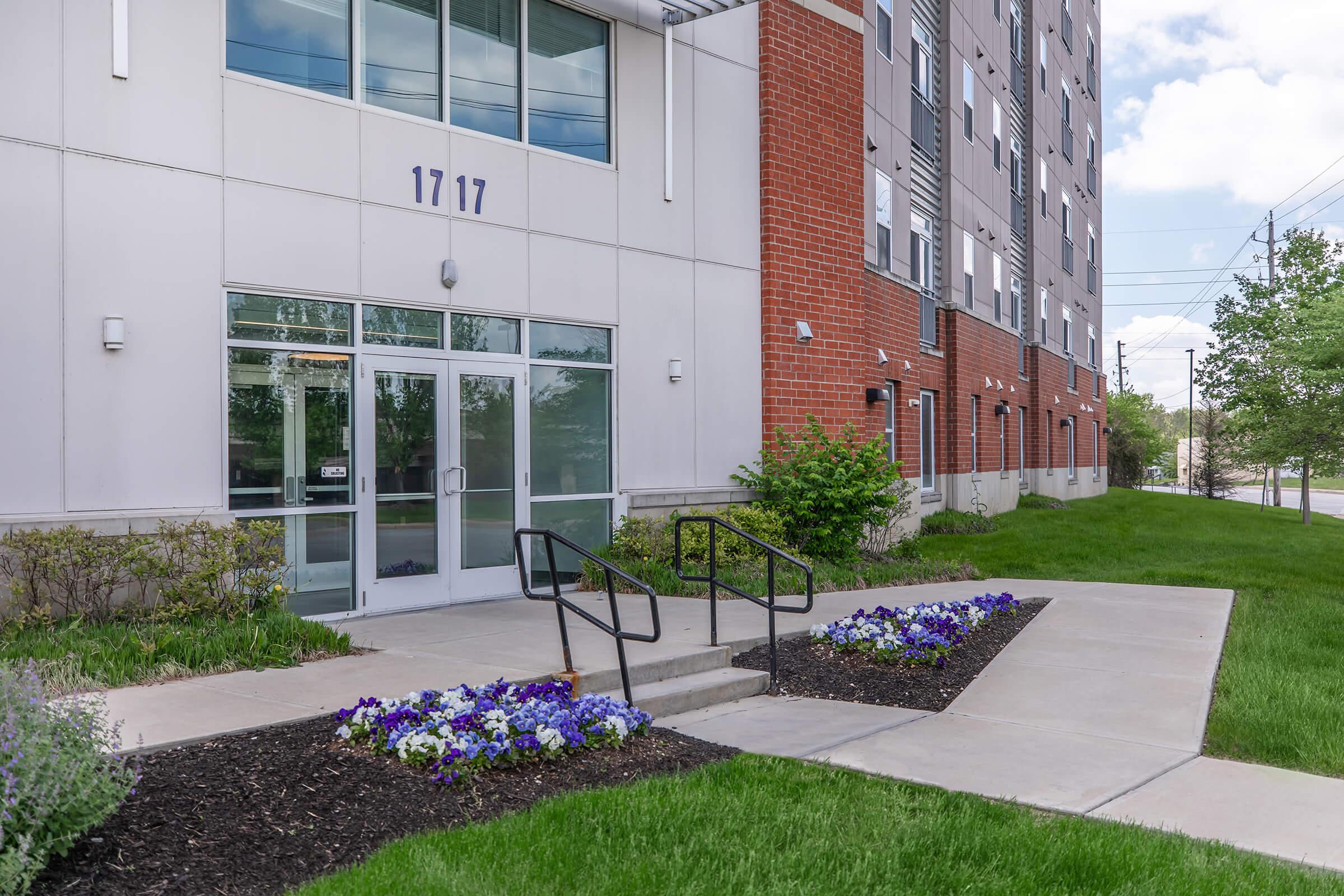 Modern building entrance featuring glass doors and a concrete pathway. Flanking the entrance are landscaped areas with green grass and flowerbeds containing purple and white flowers. There are visible railings leading to the entrance, with the building displaying the number "1717" above the door.