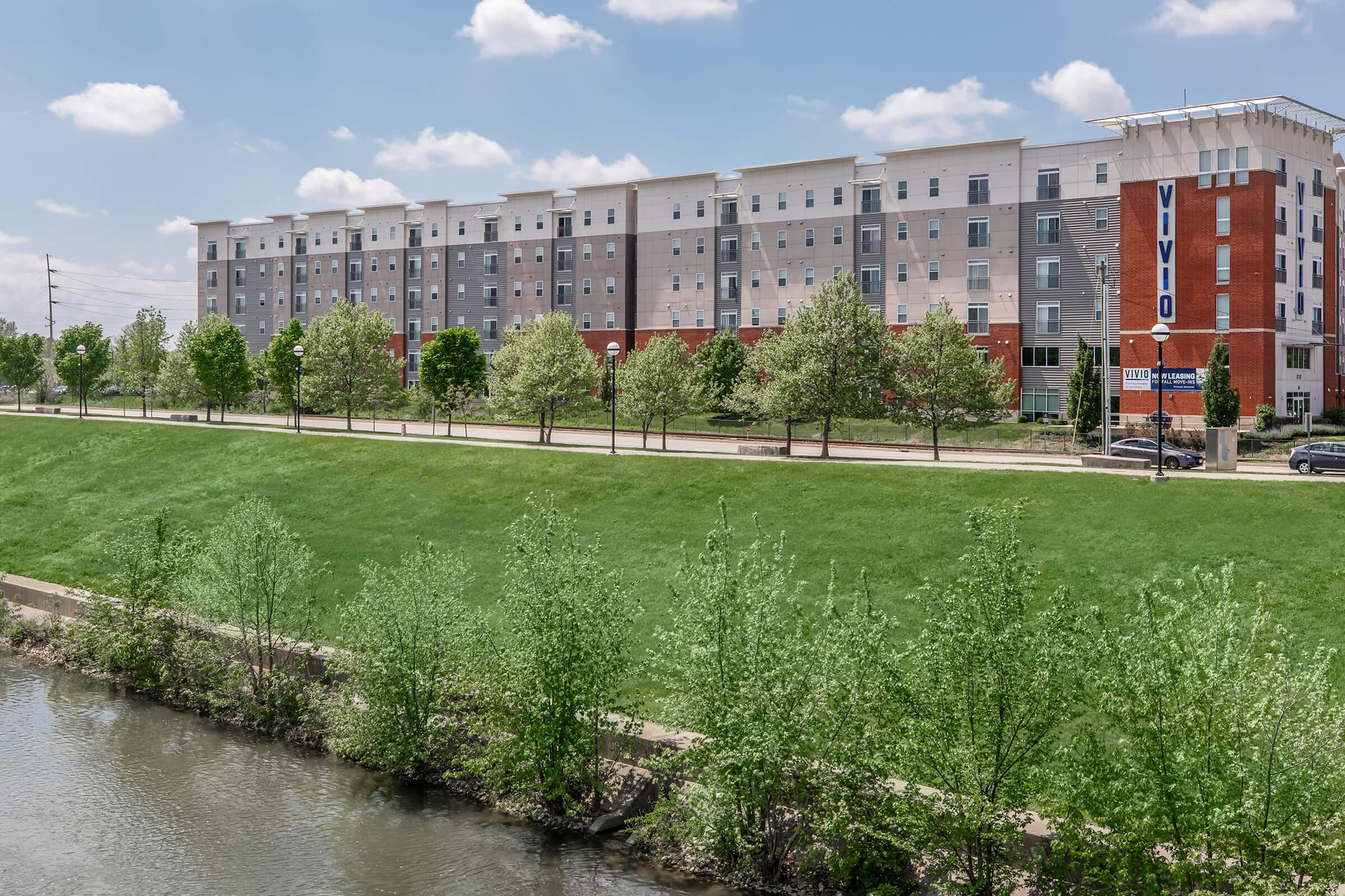 A modern multi-story residential building with a mix of brick and siding, surrounded by lush green grass and trees. A calm waterway runs in the foreground, enhancing the serene atmosphere of the area. Blue skies with fluffy clouds add to the scenic view.