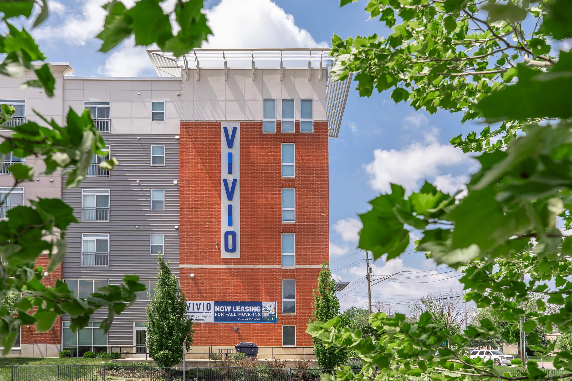 Exterior view of a modern apartment building featuring a prominent sign that reads "VIVIO" in large letters. A "NOW LEASING" banner is also displayed at the bottom. The building is surrounded by greenery and blue skies, creating a vibrant atmosphere.
