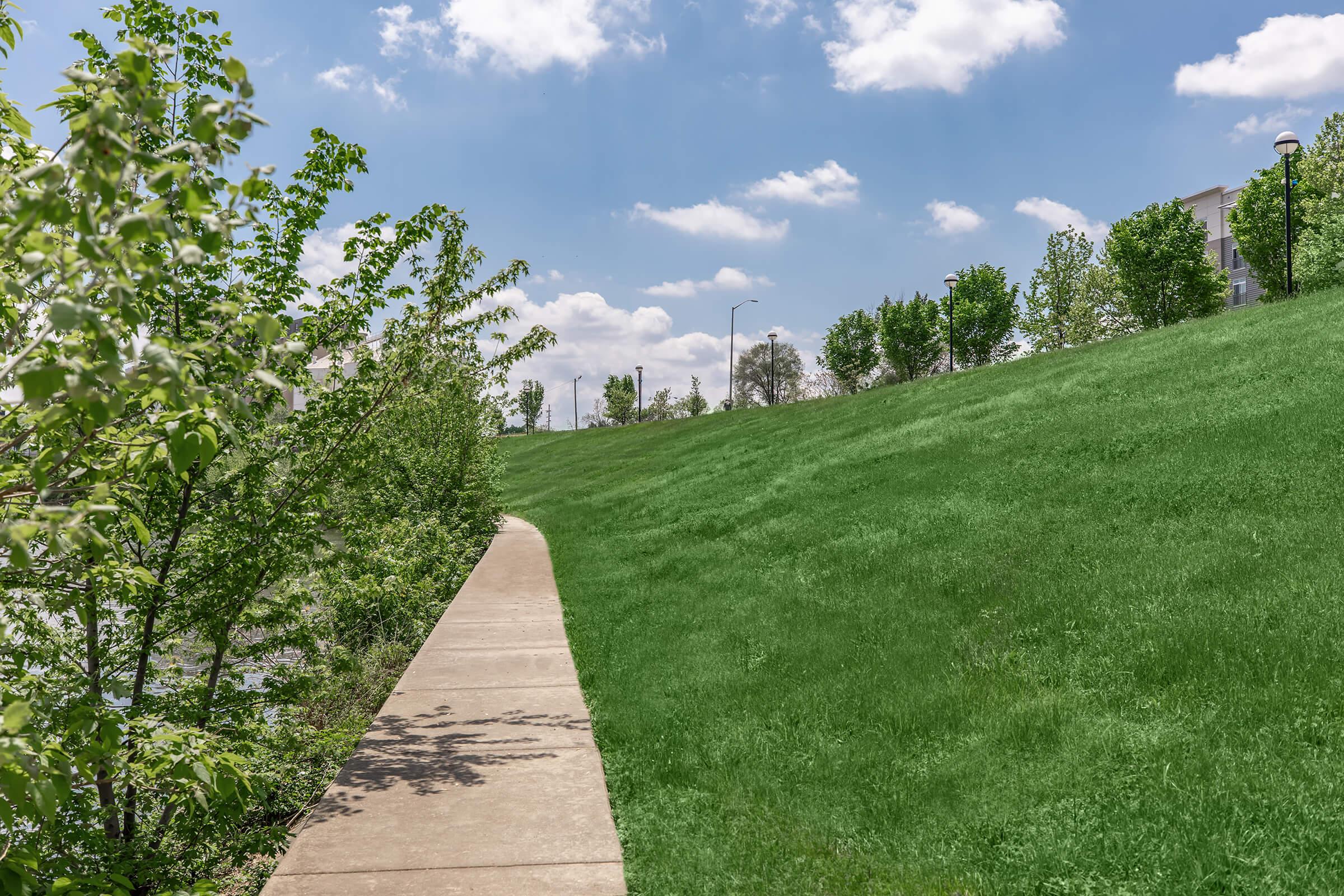 A scenic view of a winding concrete path beside a lush green hill. On the left, young trees line the pathway, while the right side features more trees in the background. The sky is bright with scattered clouds, creating a pleasant outdoor atmosphere.