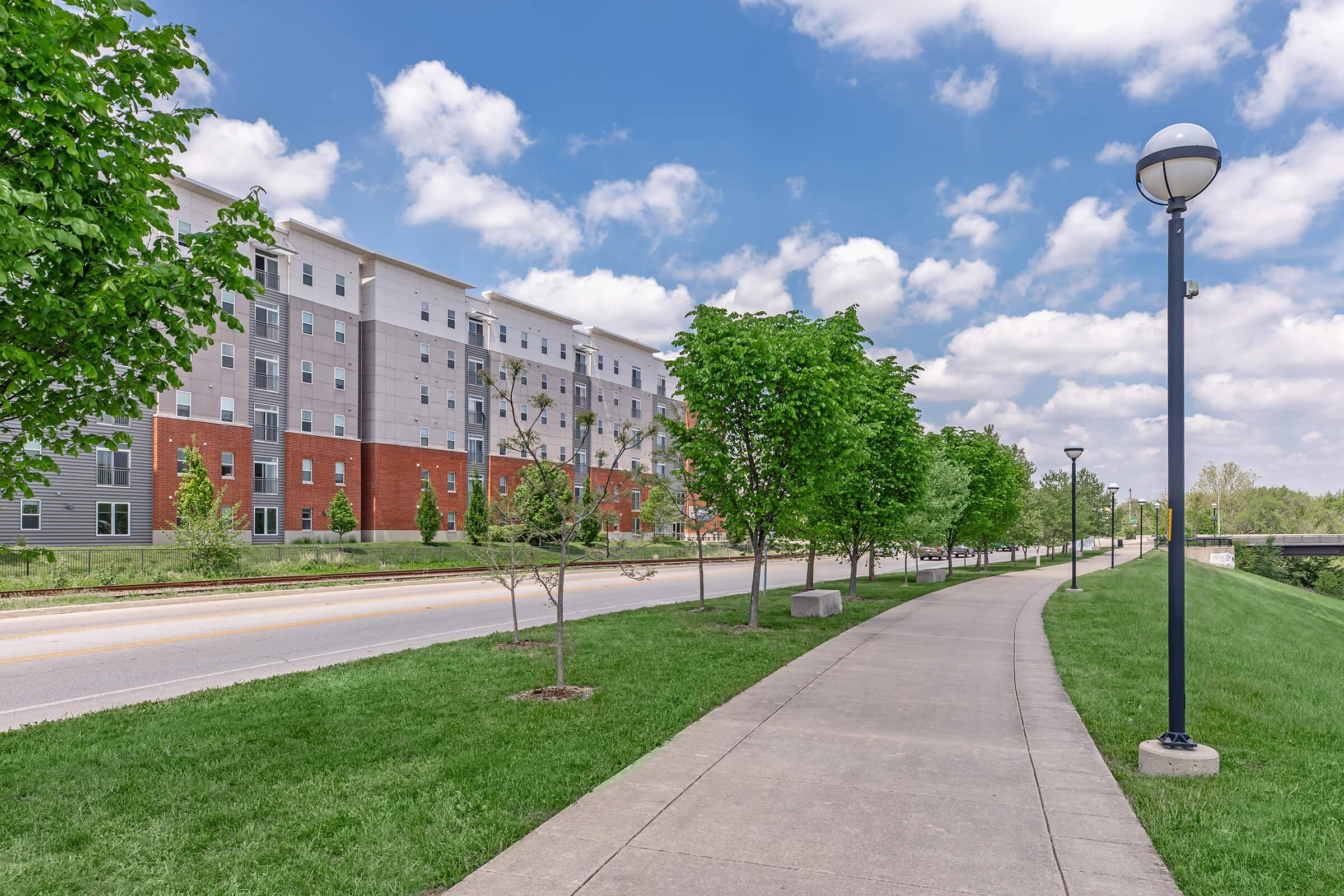 A wide sidewalk lined with green trees runs alongside a road, leading to a modern multi-story building with a mix of gray and red exterior. The sky is partly cloudy, providing a bright and inviting atmosphere. Streetlamps line the path, enhancing the urban landscape.