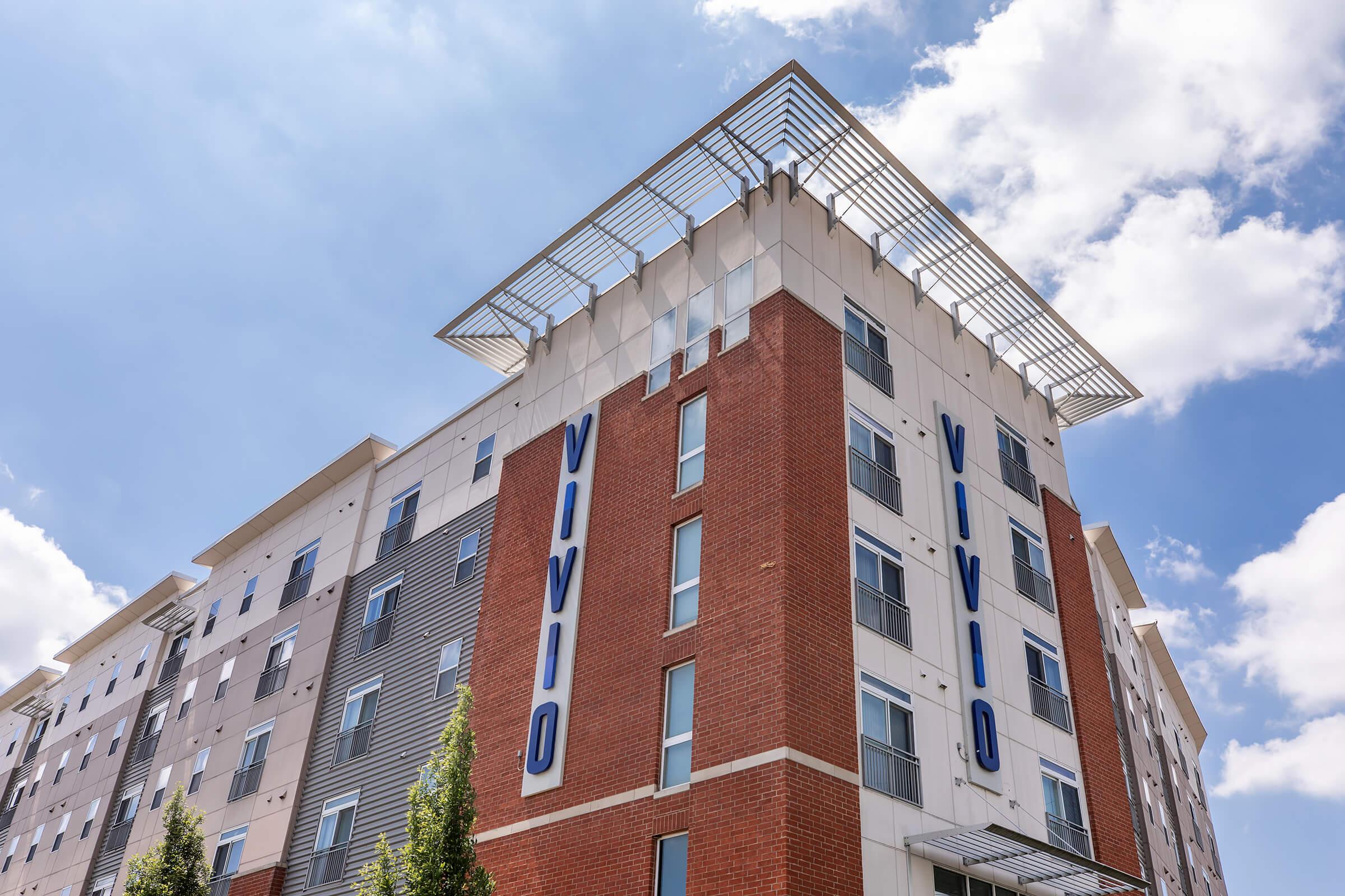 Modern apartment building with large windows and a mix of brick and metal siding. The upper section features a distinctive overhang design. Prominent "VIVIO" signage displayed on the facade. Blue sky with scattered clouds above. Green landscaping at the base adds to the inviting atmosphere.