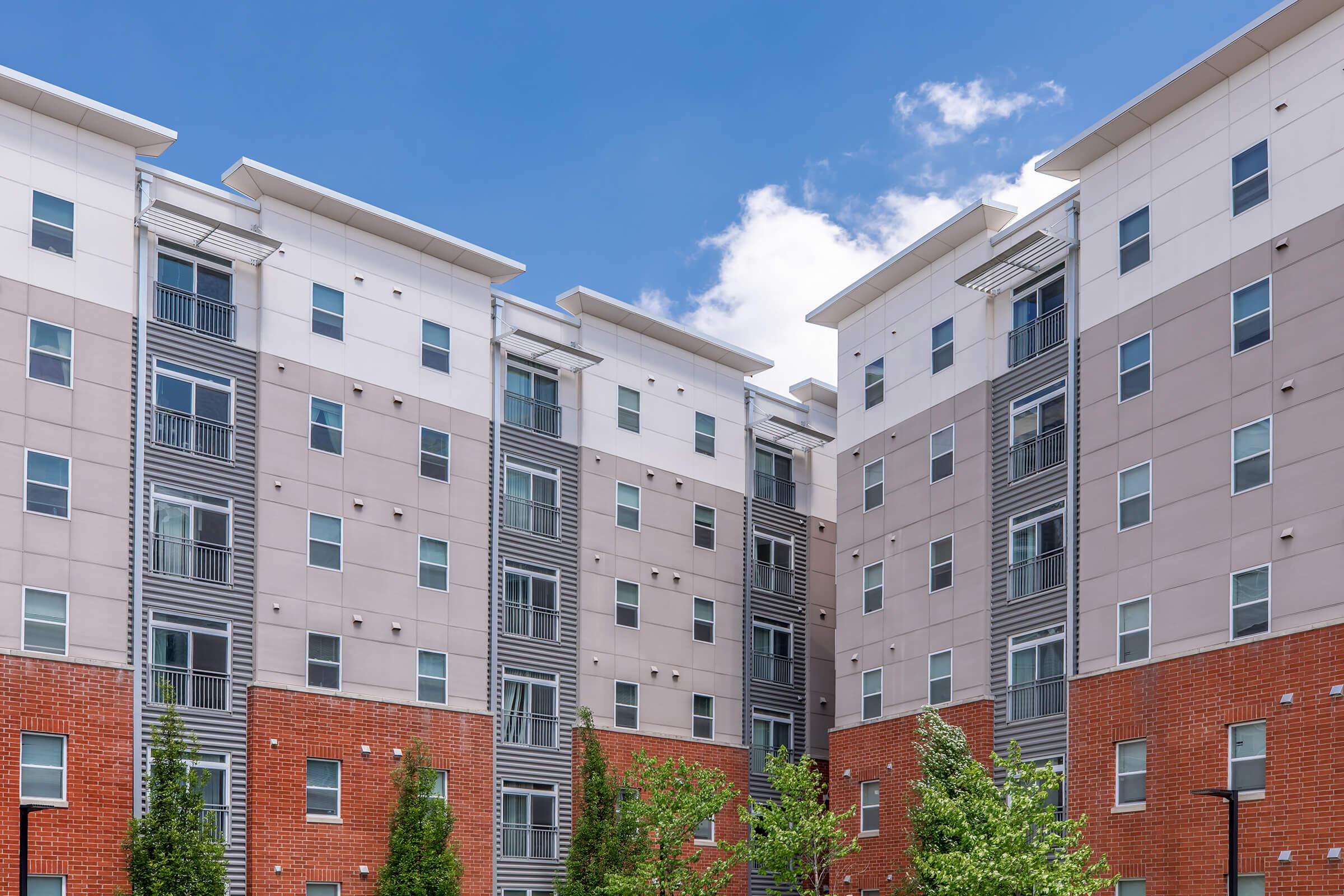 A modern apartment complex with a mix of beige and gray siding, featuring multiple levels and balconies. Trees and shrubs are planted in front, adding greenery. The sky is bright blue with a few white clouds. The overall architecture reflects contemporary design.