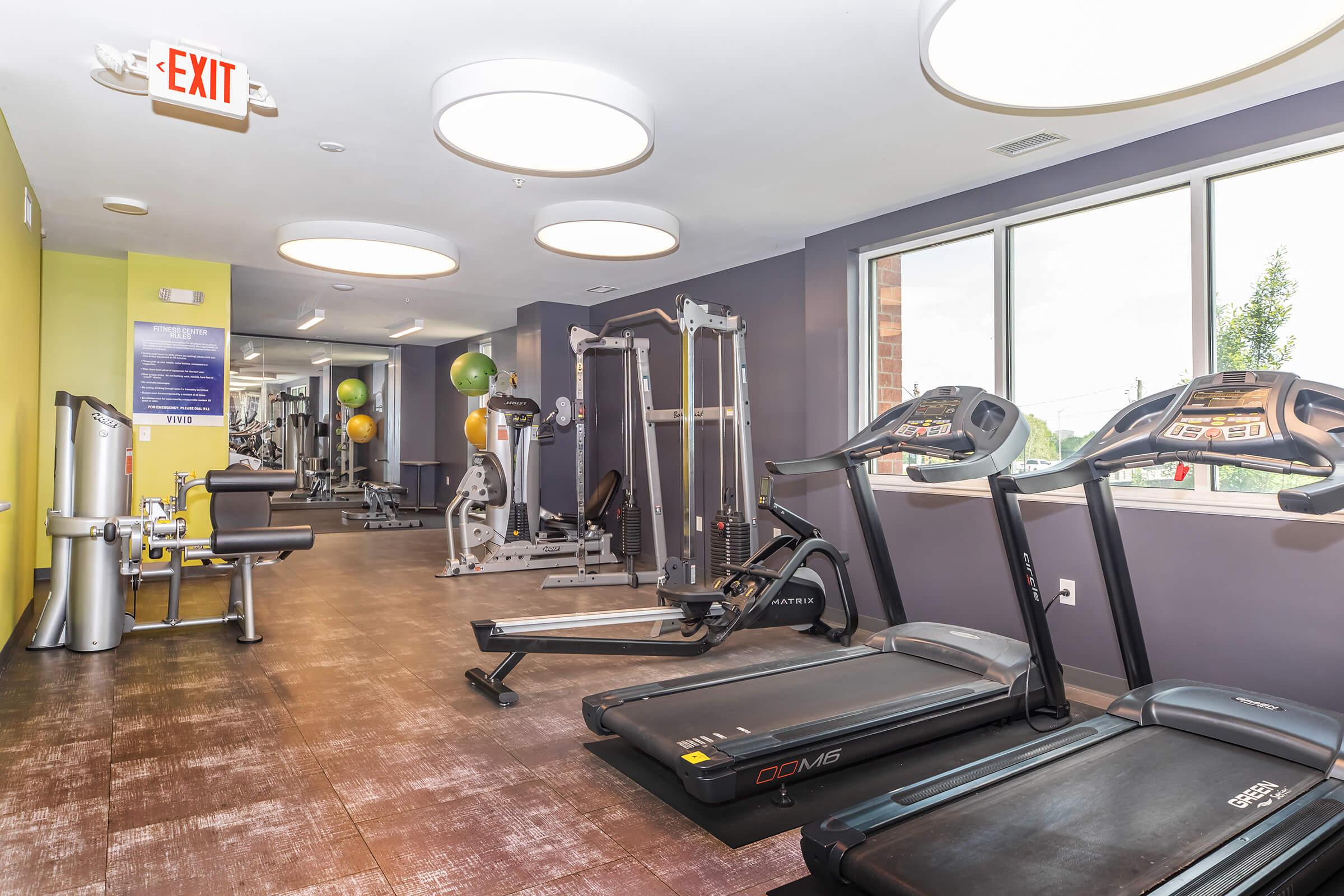 A modern gym interior featuring several exercise machines, including treadmills and weight equipment. The walls are painted in shades of purple and green, with large windows allowing natural light to enter. Exercise balls are positioned in the background, creating a welcoming fitness environment.