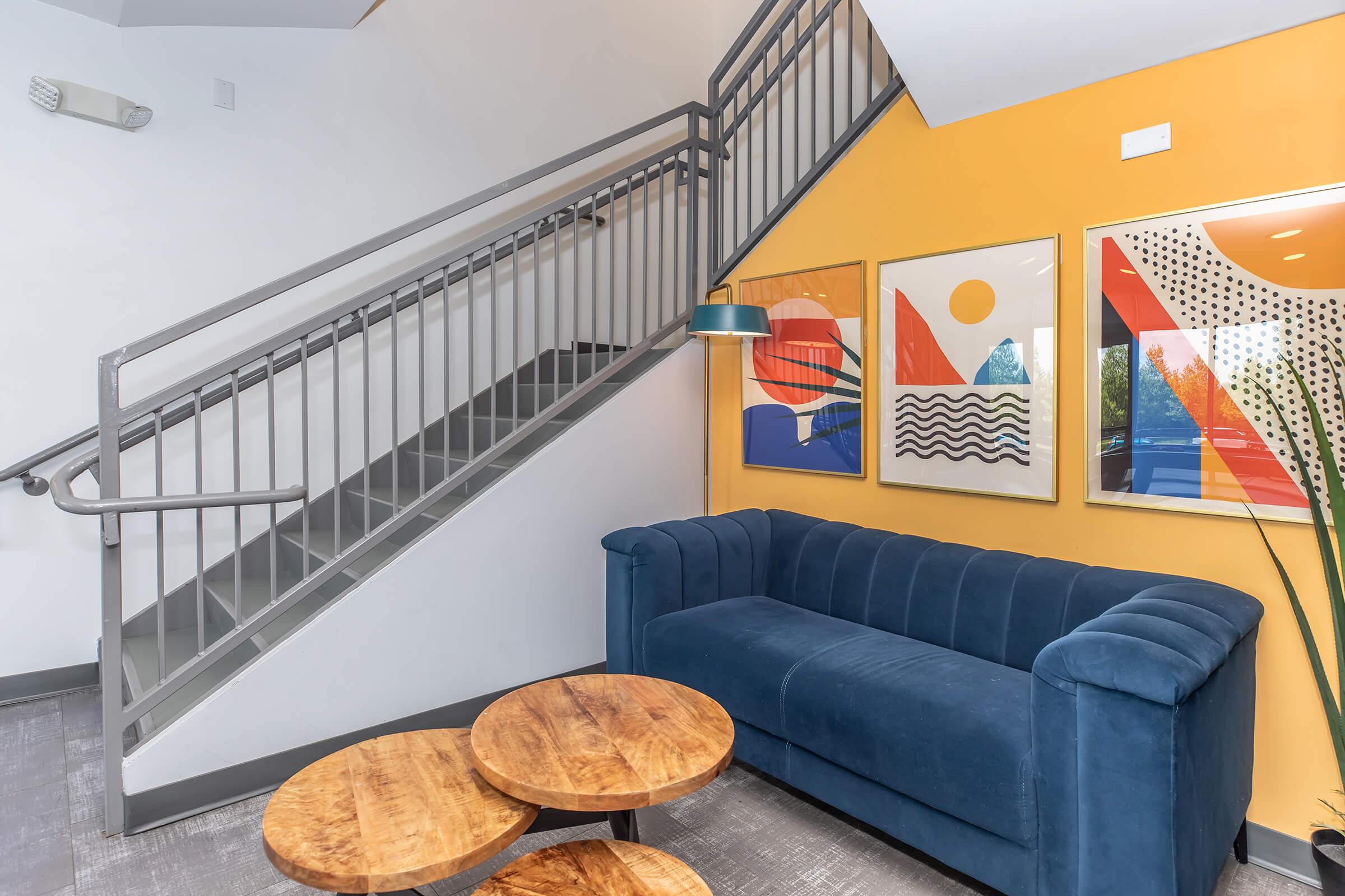 A modern interior featuring a blue velvet couch, two round wooden coffee tables, and a staircase with a metal railing. The wall is painted orange and adorned with three framed artworks, showcasing abstract designs. A lamp hangs nearby, adding a cozy atmosphere to the space.