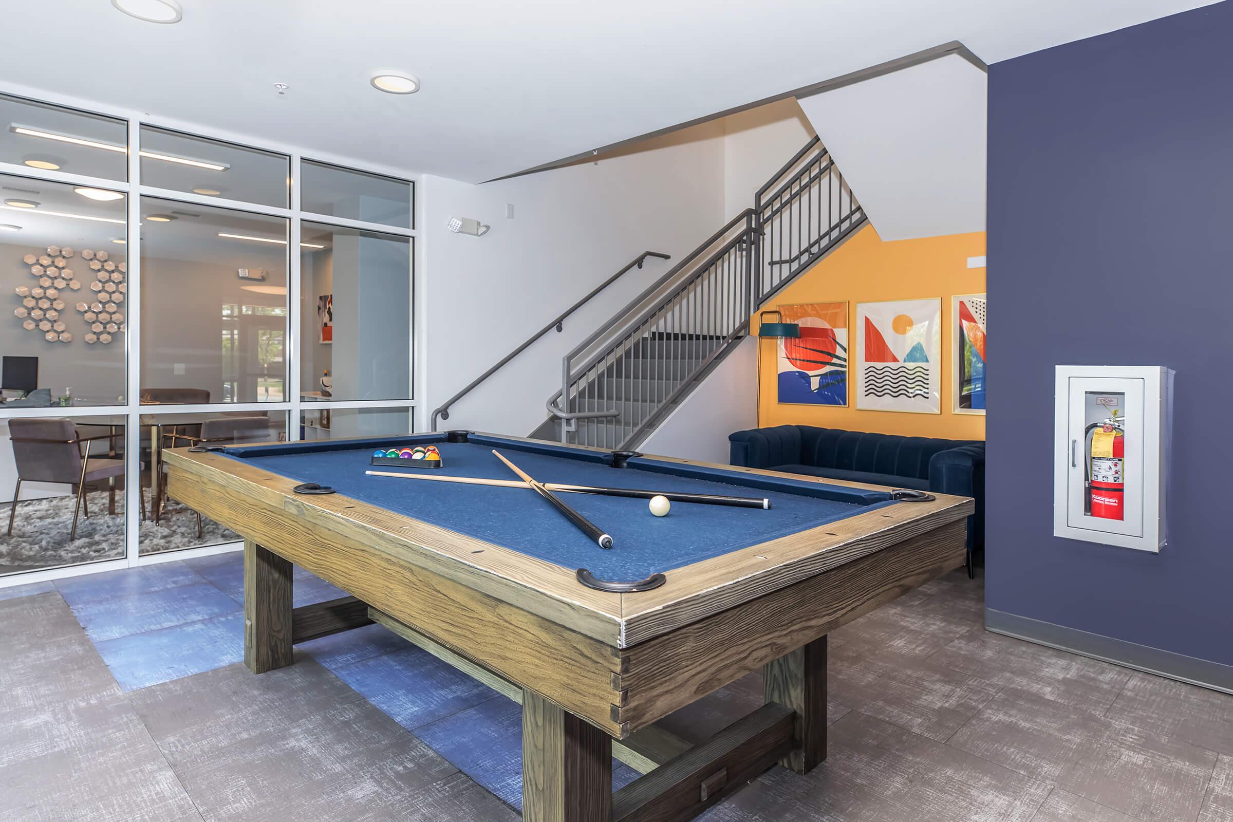 Pool table with a blue felt top, cues resting on the side, and a set of pool balls. The room features a staircase in the background, colorful wall art, and a blue couch. A fire extinguisher is mounted on a wall nearby. The space is well-lit and designed for recreational use.