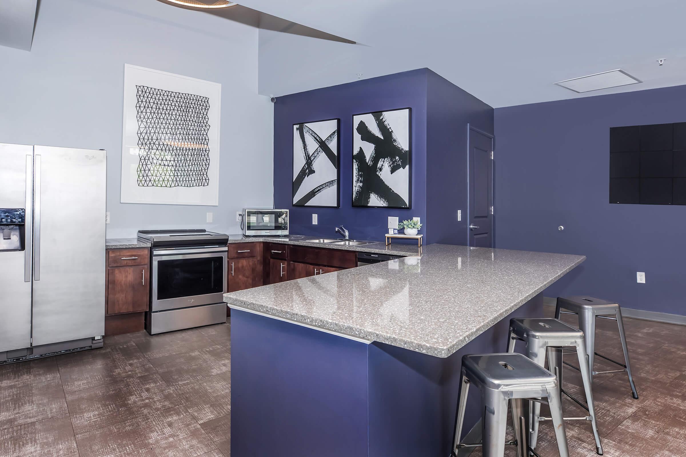 Modern kitchen featuring dark blue walls, stainless steel appliances, and a granite countertop. The space includes a silver refrigerator, oven, and microwave, complemented by metal stools at the counter. Two abstract framed artworks are displayed on the wall.