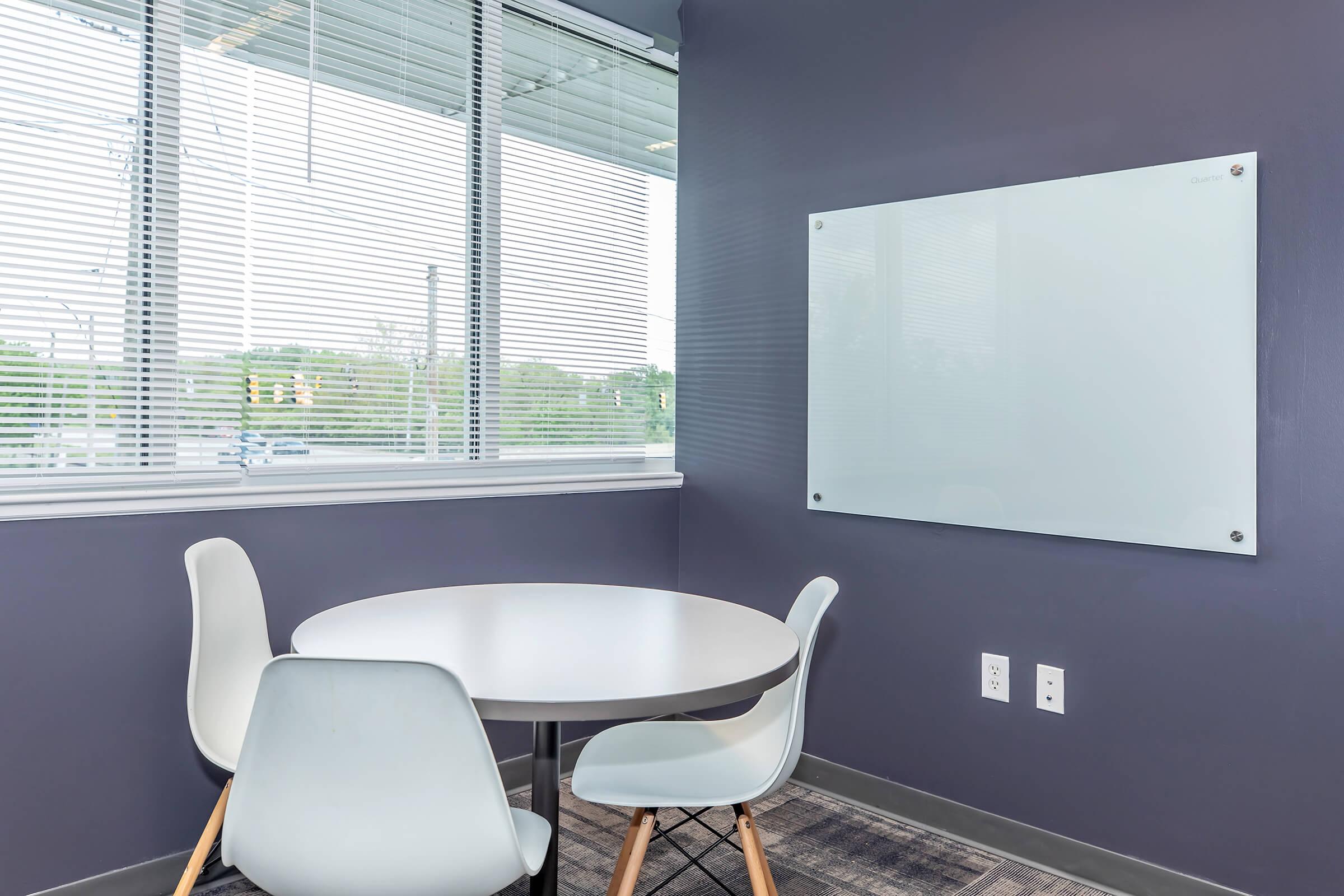 A modern office space featuring a small round table with three chairs. The walls are painted in a dark color, and there is a large whiteboard mounted on the wall. Natural light filters in through large windows with blinds, and there are green trees visible outside.