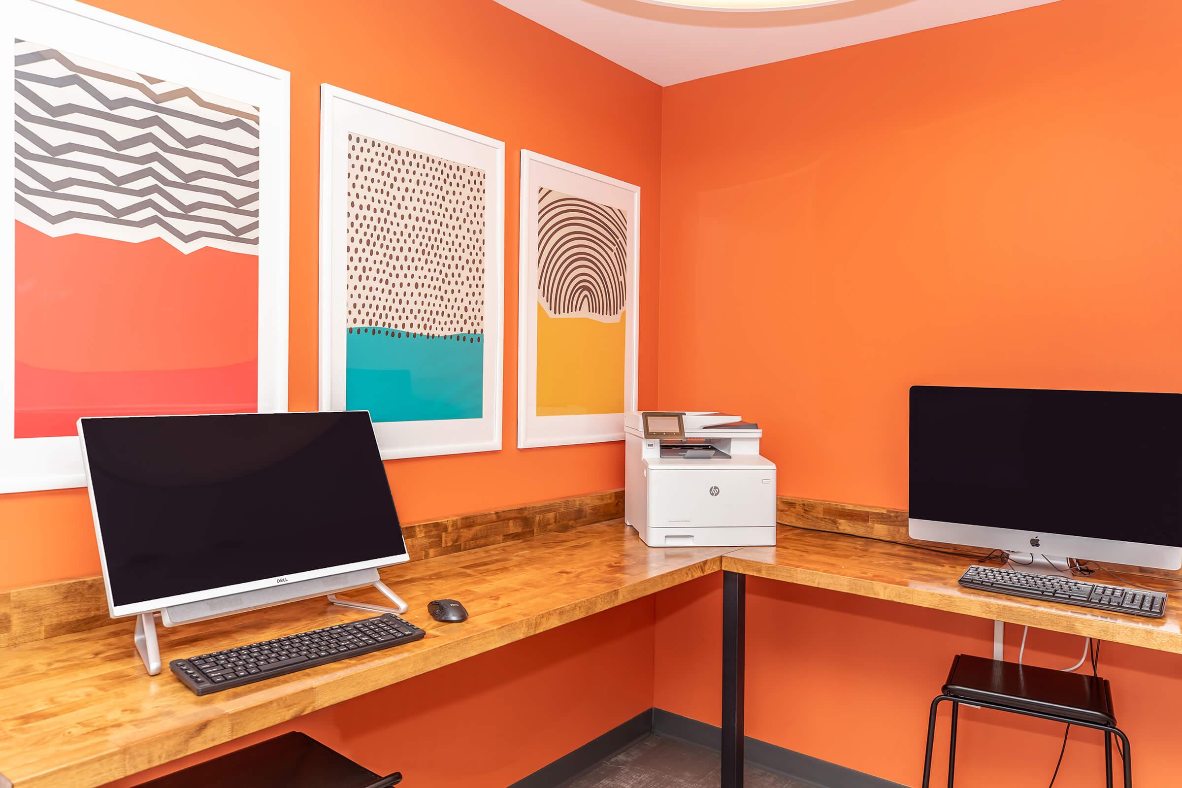 A bright coworking space with an orange wall, featuring three abstract art prints. There are two desktop computers with keyboards on a wooden L-shaped desk, and a printer beside them. The modern setup provides a productive atmosphere for work or collaboration.