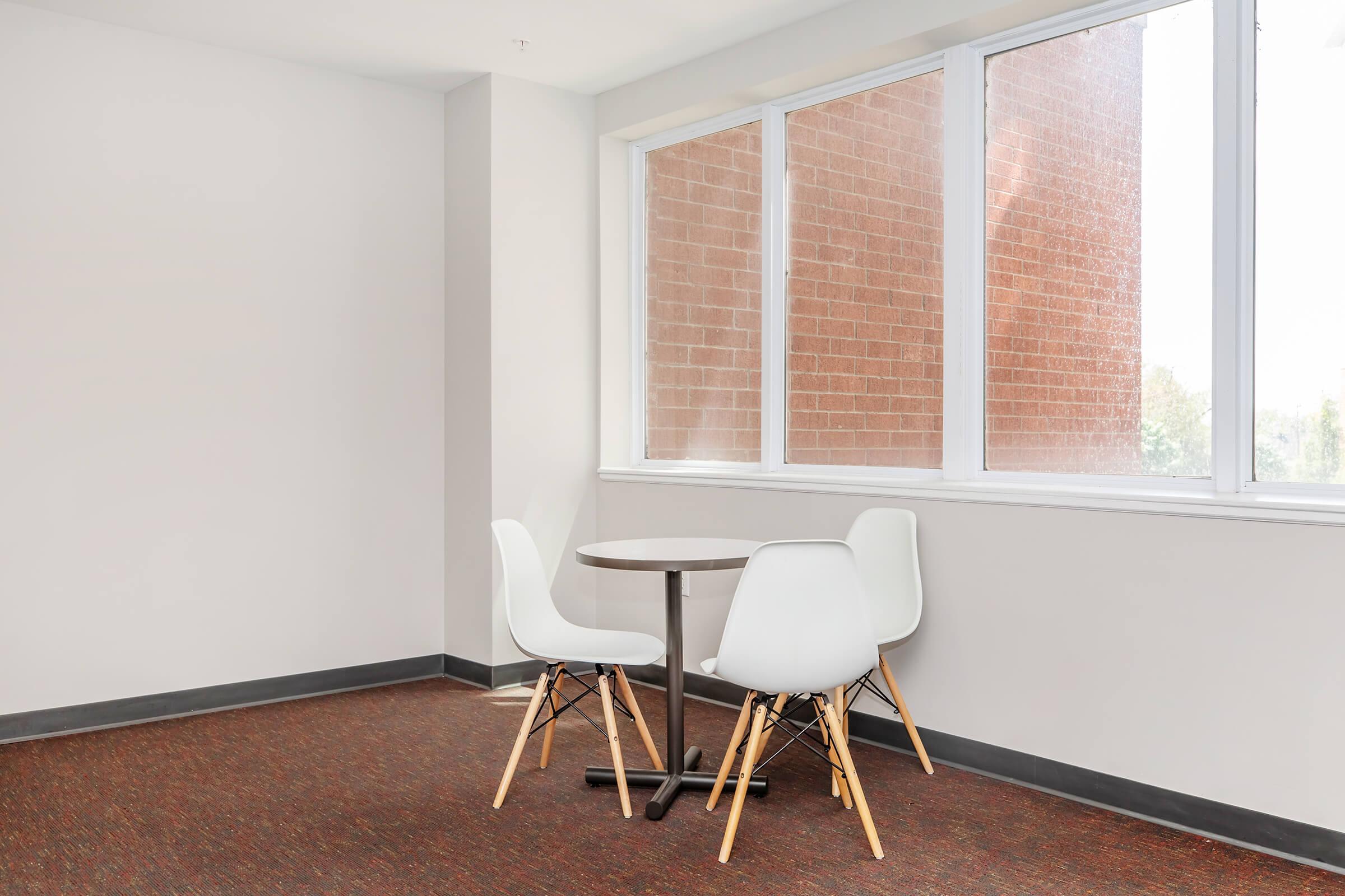 A small seating area featuring a round table with two modern white chairs, situated near a large window that overlooks a brick wall. The room has light-colored walls and a textured brown carpet.