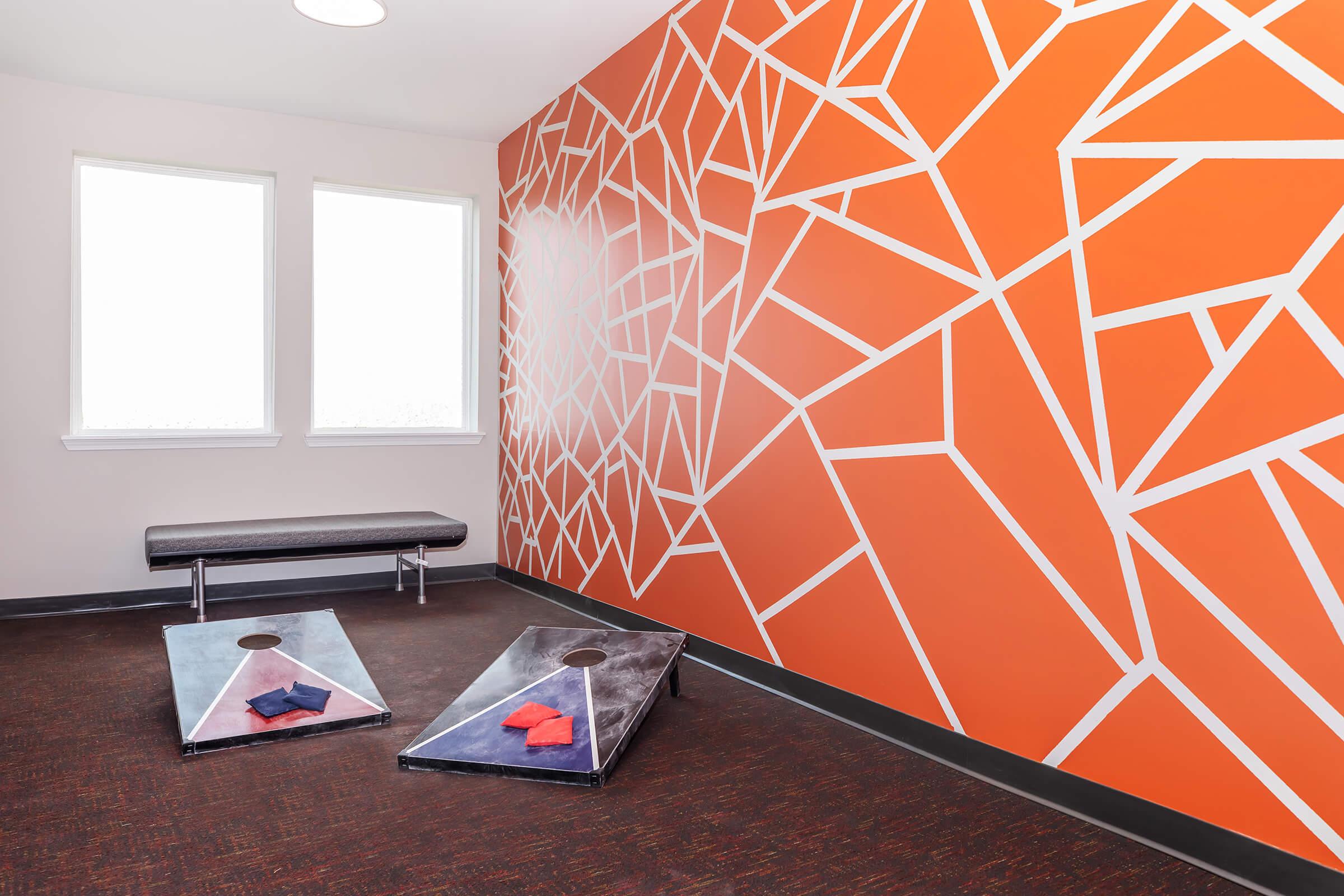 A modern recreational space featuring a vibrant orange geometric wall, two cornhole boards with red and blue bags, and a sleek bench. Natural light comes through the windows, creating a welcoming atmosphere for entertainment and relaxation.