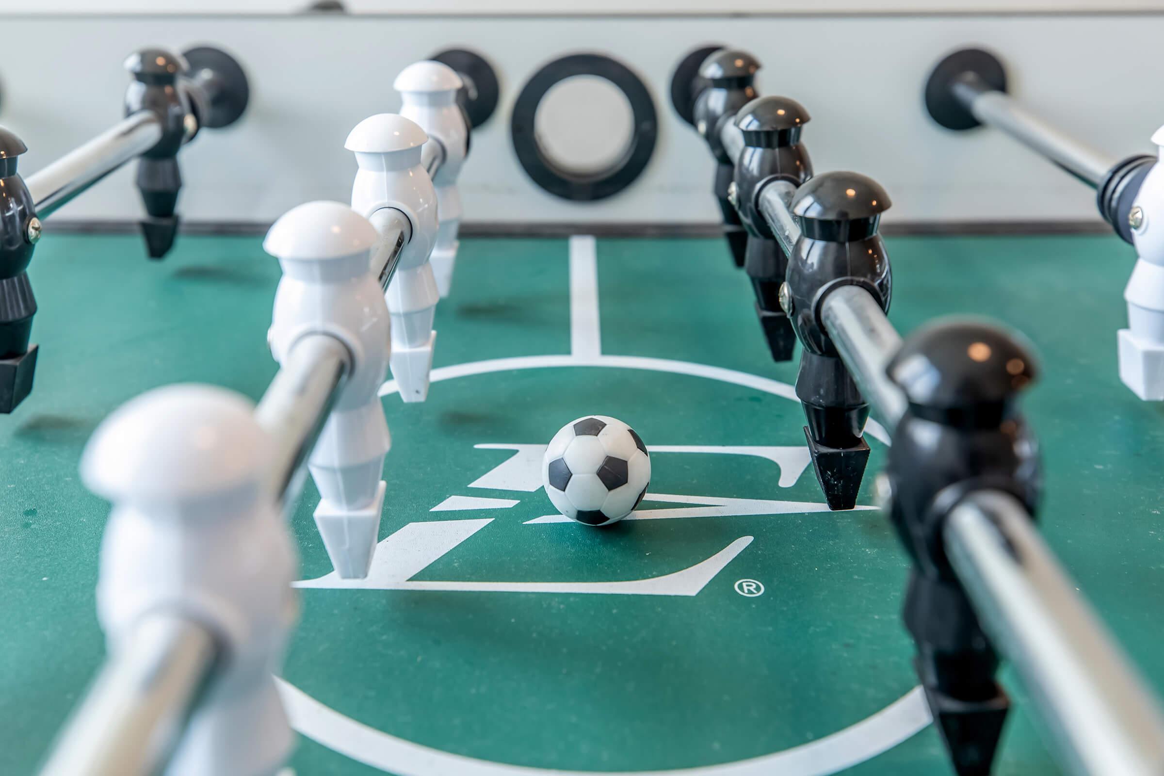A close-up view of a foosball table featuring two rows of black and white player figures. A small soccer ball is positioned in the center of the table, with a logo visible in the background on the green tabletop. The scene captures the excitement of a game in action.