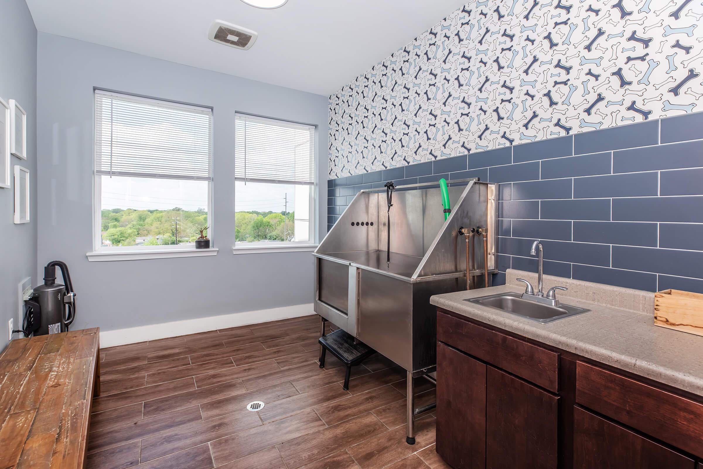 A spacious pet grooming room featuring a stainless steel tub with adjustable sprayers, a wall-mounted sink, and wooden countertops. The walls are decorated with a playful dog bone pattern, while large windows provide natural light and a view of the outdoors. The floor is made of dark, wooden tiles.
