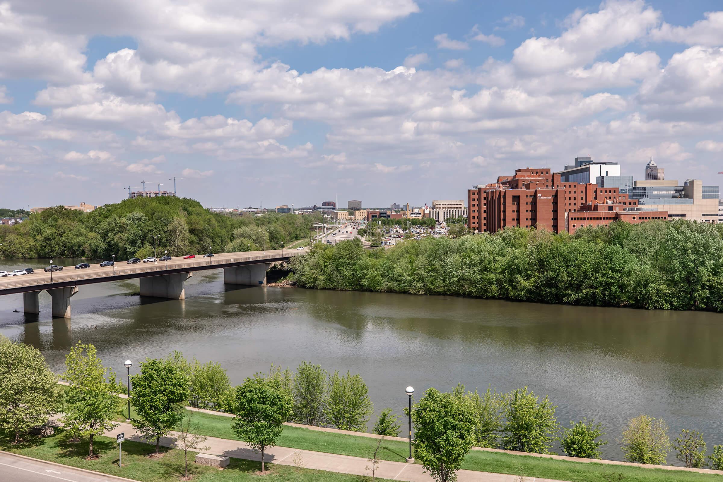 A scenic view of a river with a bridge crossing over it, surrounded by lush green trees. In the background, a city skyline with various buildings under a partly cloudy sky can be seen. The setting suggests a peaceful urban environment.