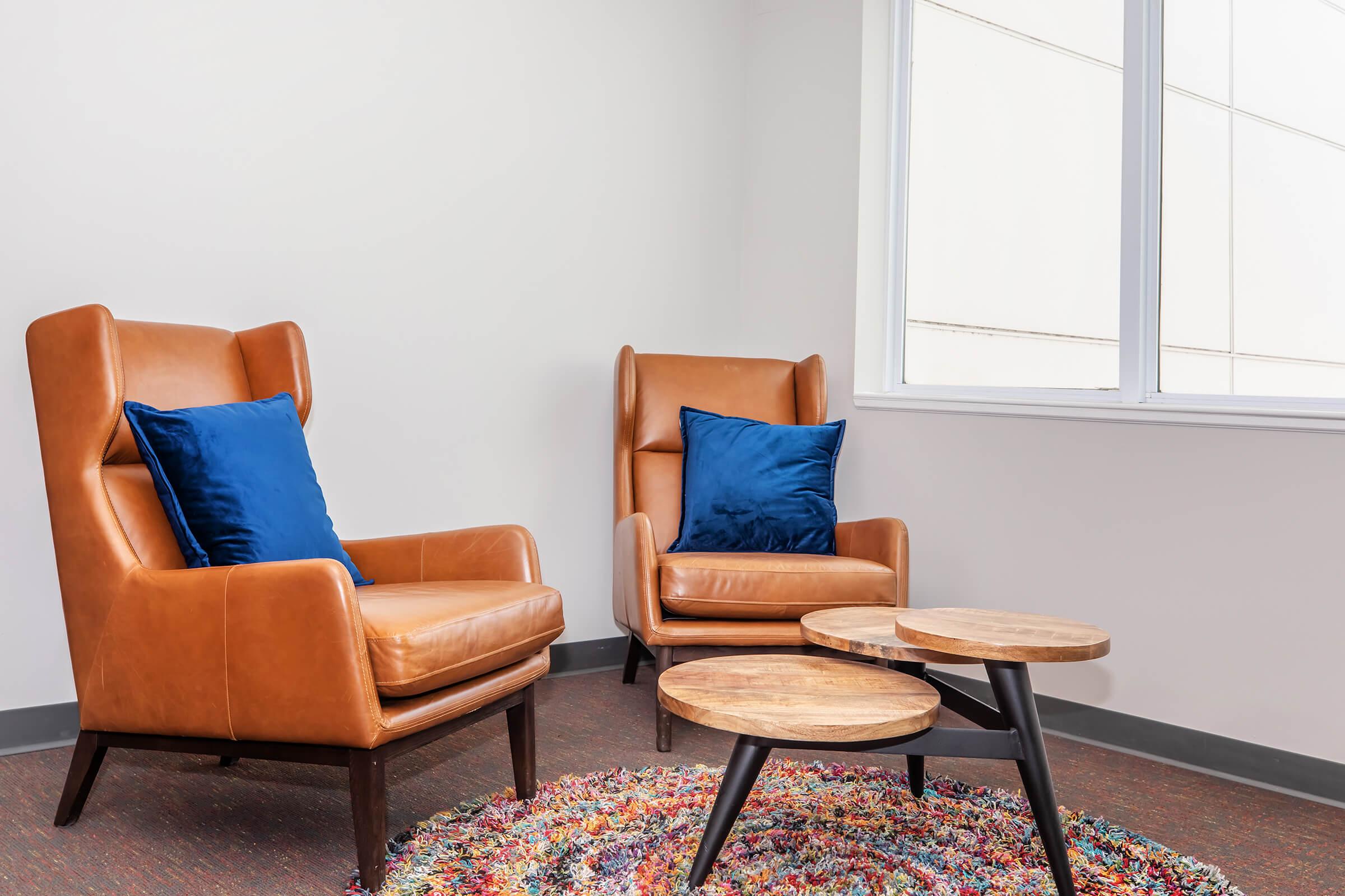 A cozy seating area featuring two brown leather armchairs with blue cushions, positioned beside a stylish wooden coffee table. A colorful, textured rug lies on the floor, adding warmth to the modern, minimalist space with a large window allowing natural light.
