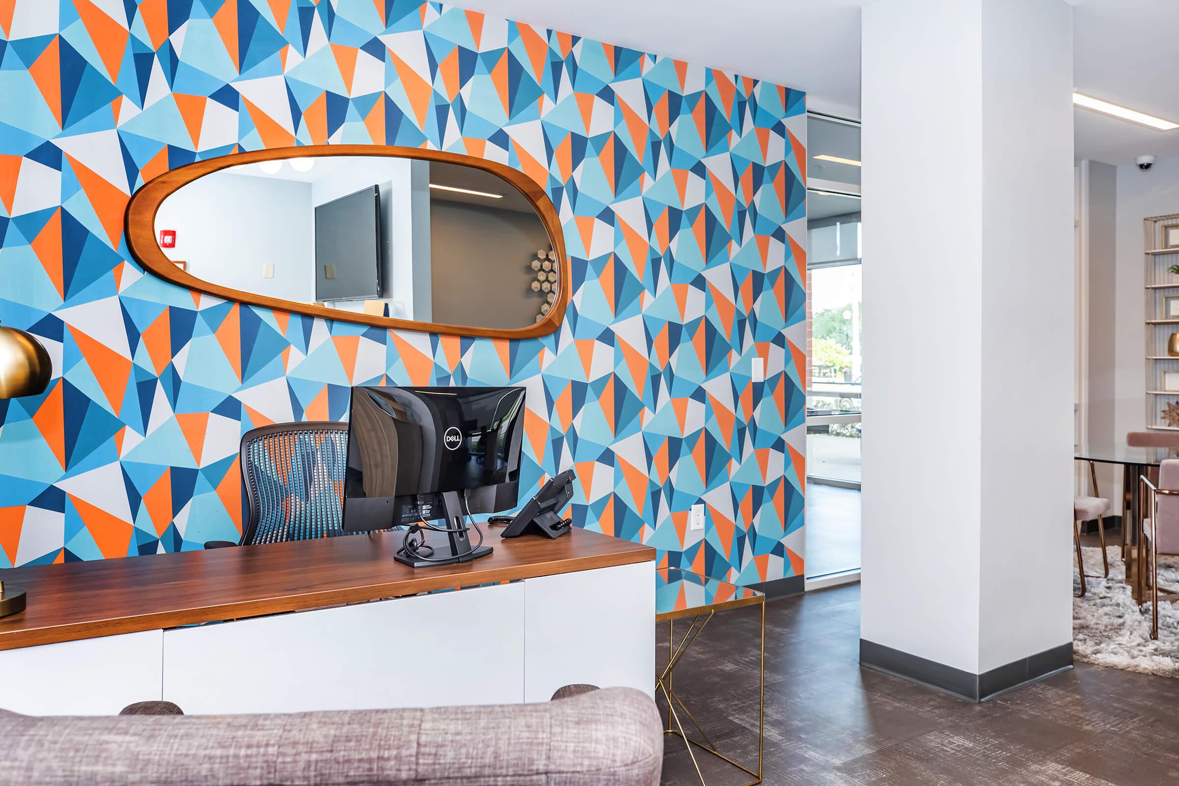 A modern office space featuring a colorful geometric patterned accent wall in blue, orange, and white. A sleek desk with a computer monitor and telephone is positioned in front of the wall. A round mirror above the desk reflects the vibrant design, while a comfortable chair and a cozy seating area are visible in the background.