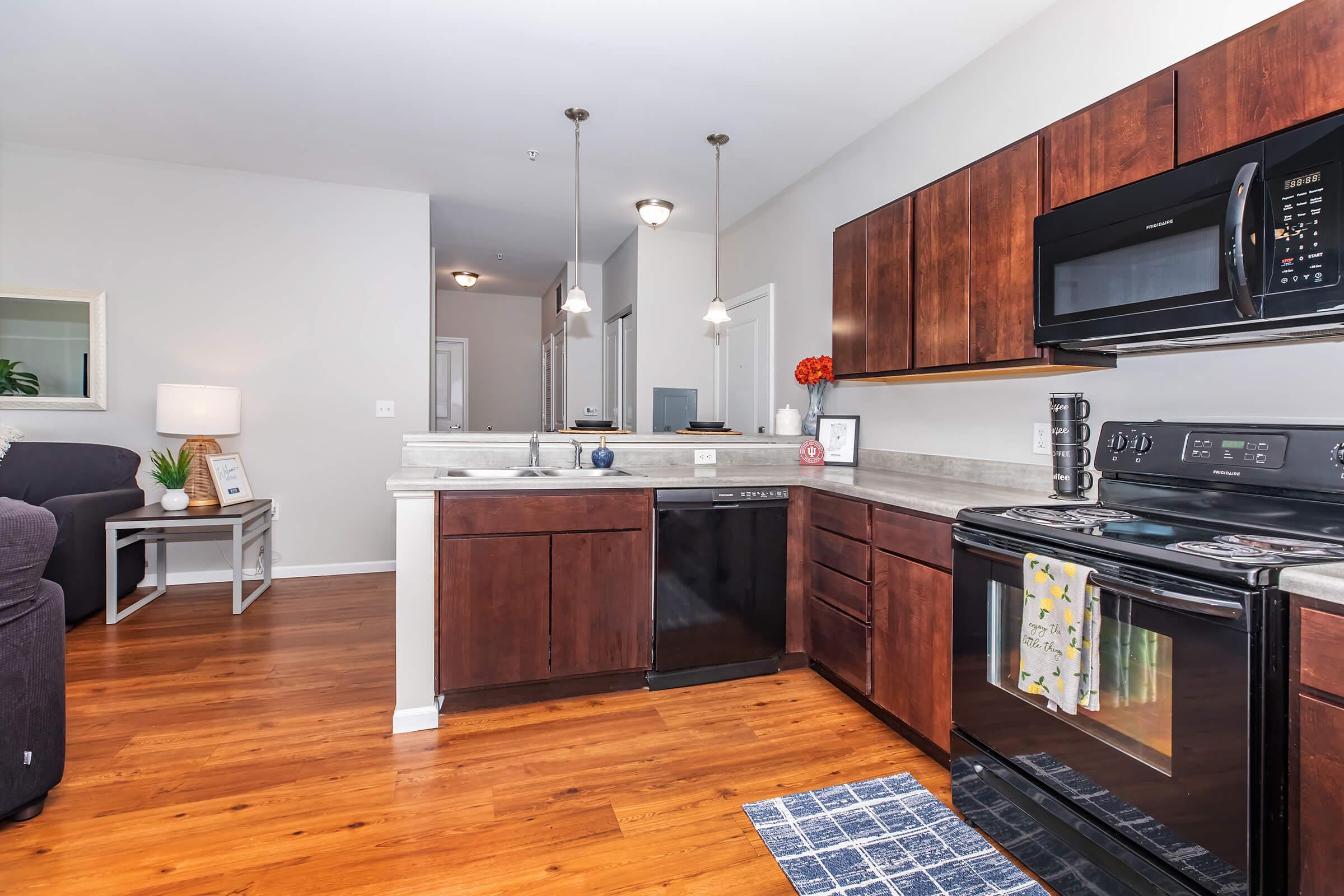 Modern kitchen with dark wooden cabinets, stainless steel appliances, and a gray countertop. Includes a microwave, oven, dishwasher, and sink, with decorative items like kitchen towels and flowers. The living area is visible in the background, featuring a lamp and a chair.