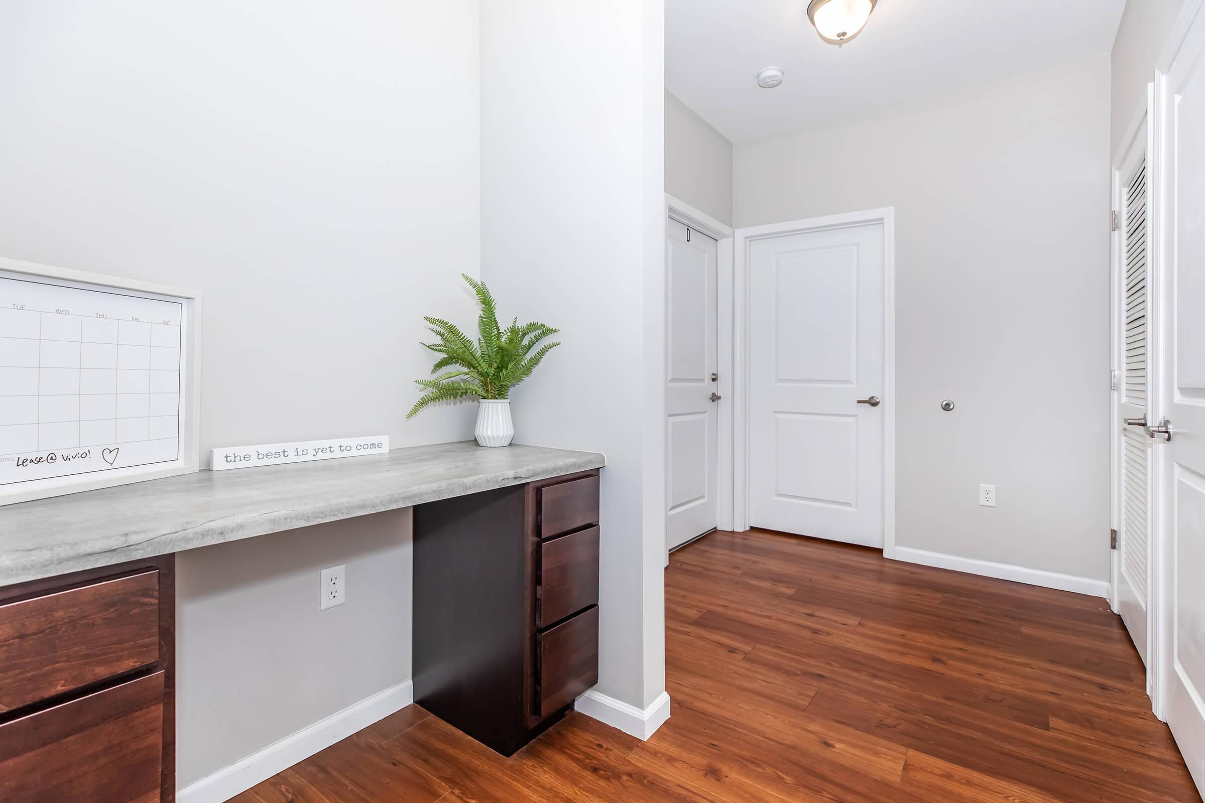 Modern interior with a small desk and drawers against a light gray wall. A potted plant adds a touch of green beside the desk, which features a sign with motivational text. The space has wooden flooring and a bright, open feel with a door leading to another room.