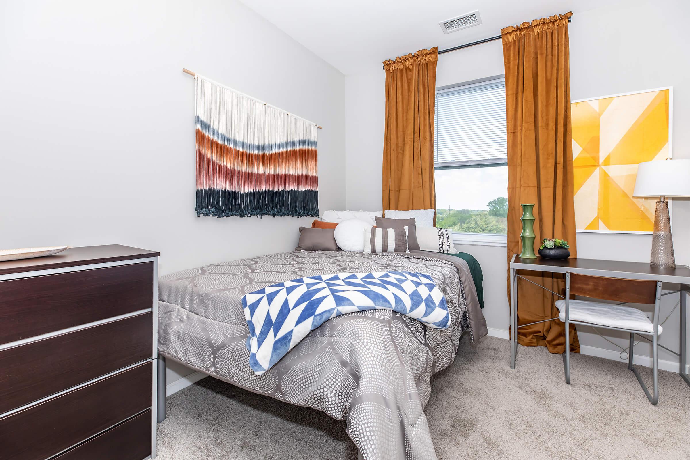 A cozy bedroom featuring a queen-sized bed with decorative pillows and a textured blanket. There's a wooden dresser, a small desk with a lamp and orange curtains, and wall decor including a tapestry and an abstract painting in vibrant colors. The room has a warm and inviting atmosphere.