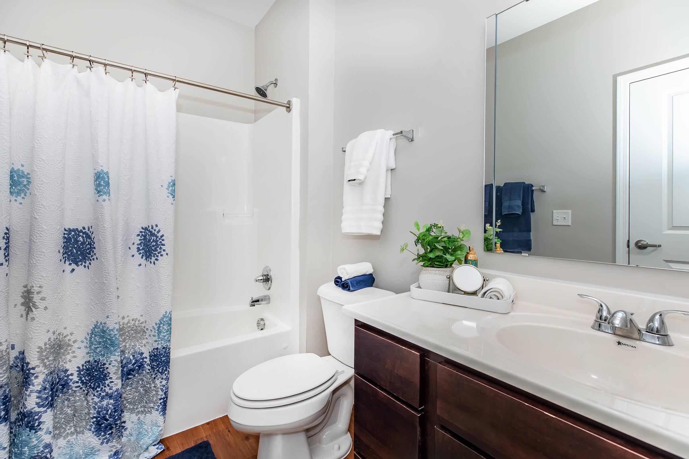A well-lit bathroom featuring a white shower curtain with blue and gray patterns, a bathtub with a glass sliding door, a white toilet, and a sink with dark wood cabinetry. The countertop is neatly arranged with towels and a small potted plant. A large mirror and a towel rack are visible.