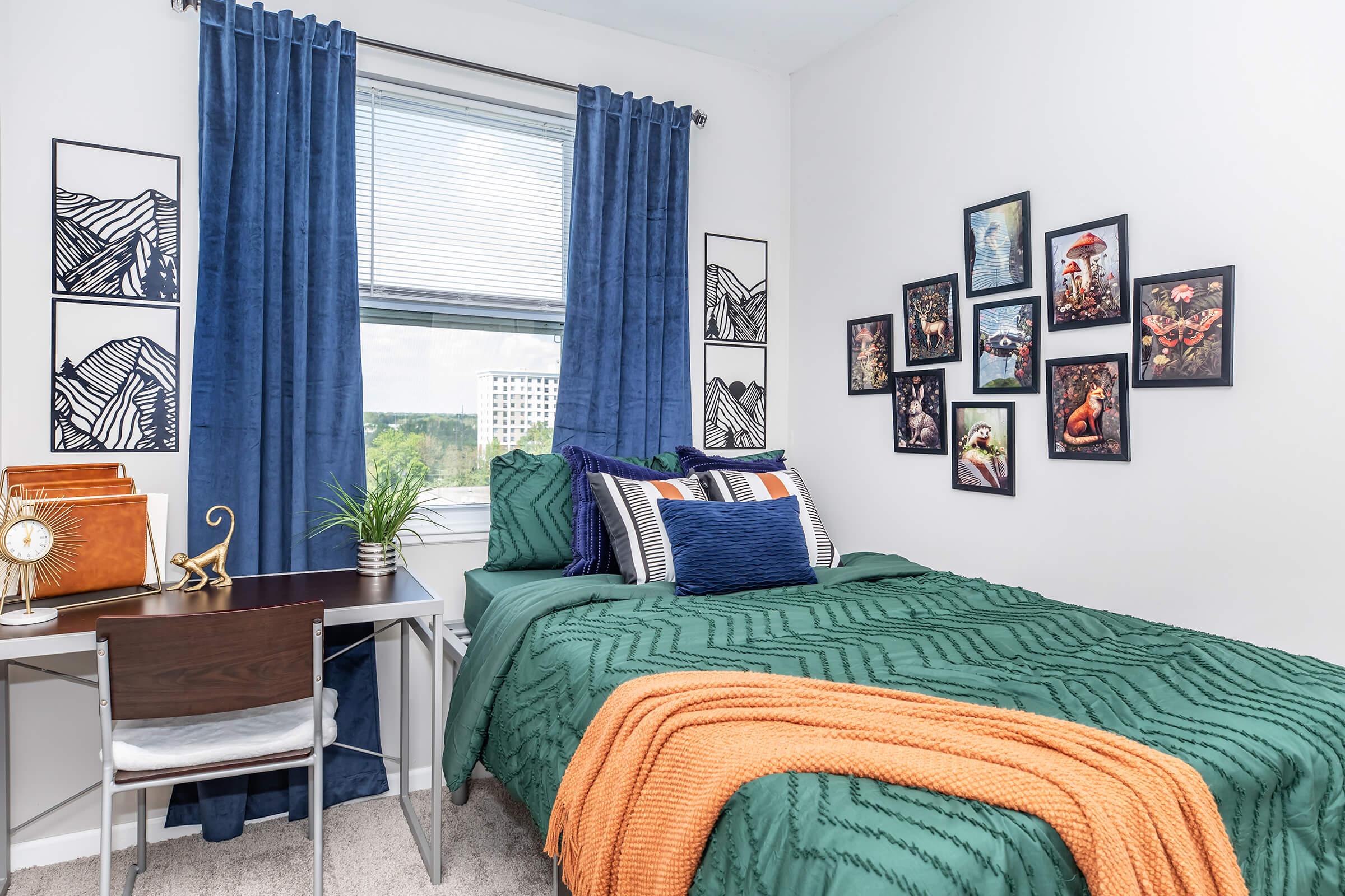 A cozy bedroom featuring a neatly made bed with green bedding and colorful pillows, a desk with a small plant, and a window with blue curtains. The walls are decorated with framed artwork, and the overall design has a modern and inviting atmosphere.