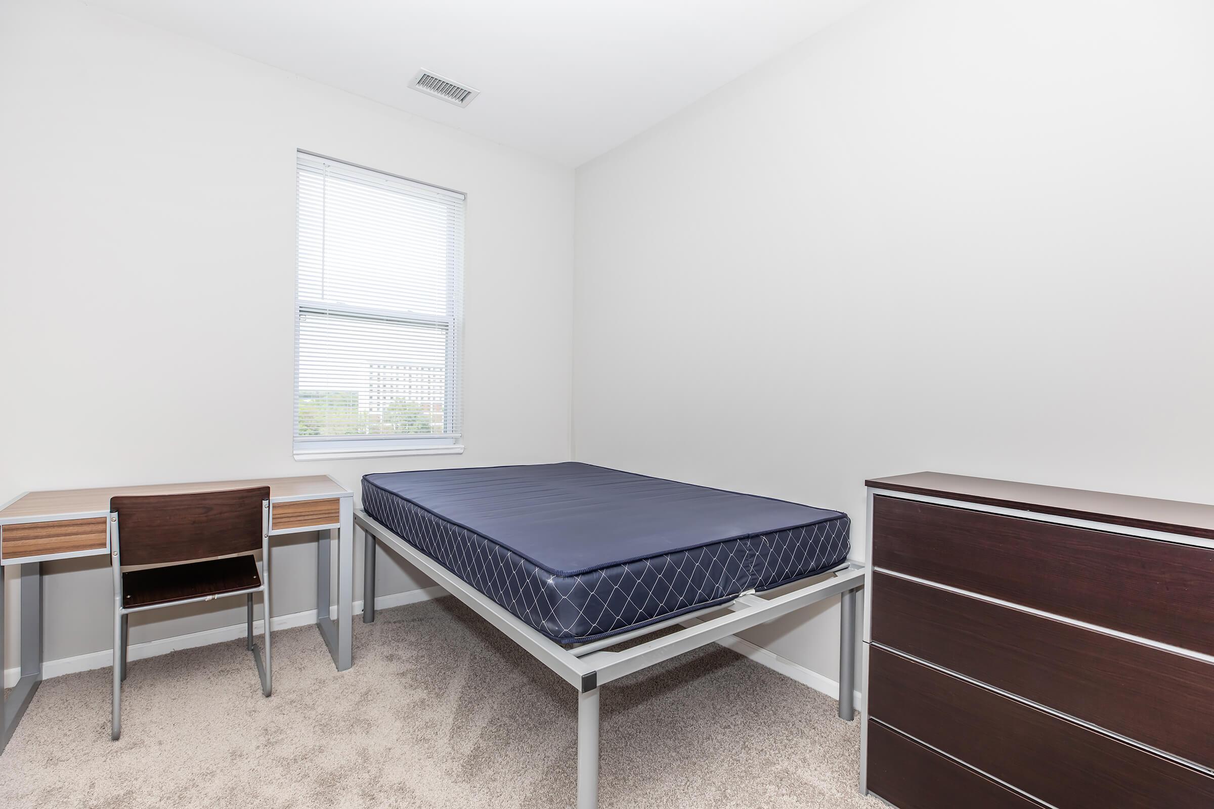 A minimalistic bedroom featuring a single bed with a blue mattress, a wooden desk and chair, and a dark wood dresser. The walls are painted white, and there is a window allowing natural light to enter the room. The flooring is carpeted in a neutral tone.