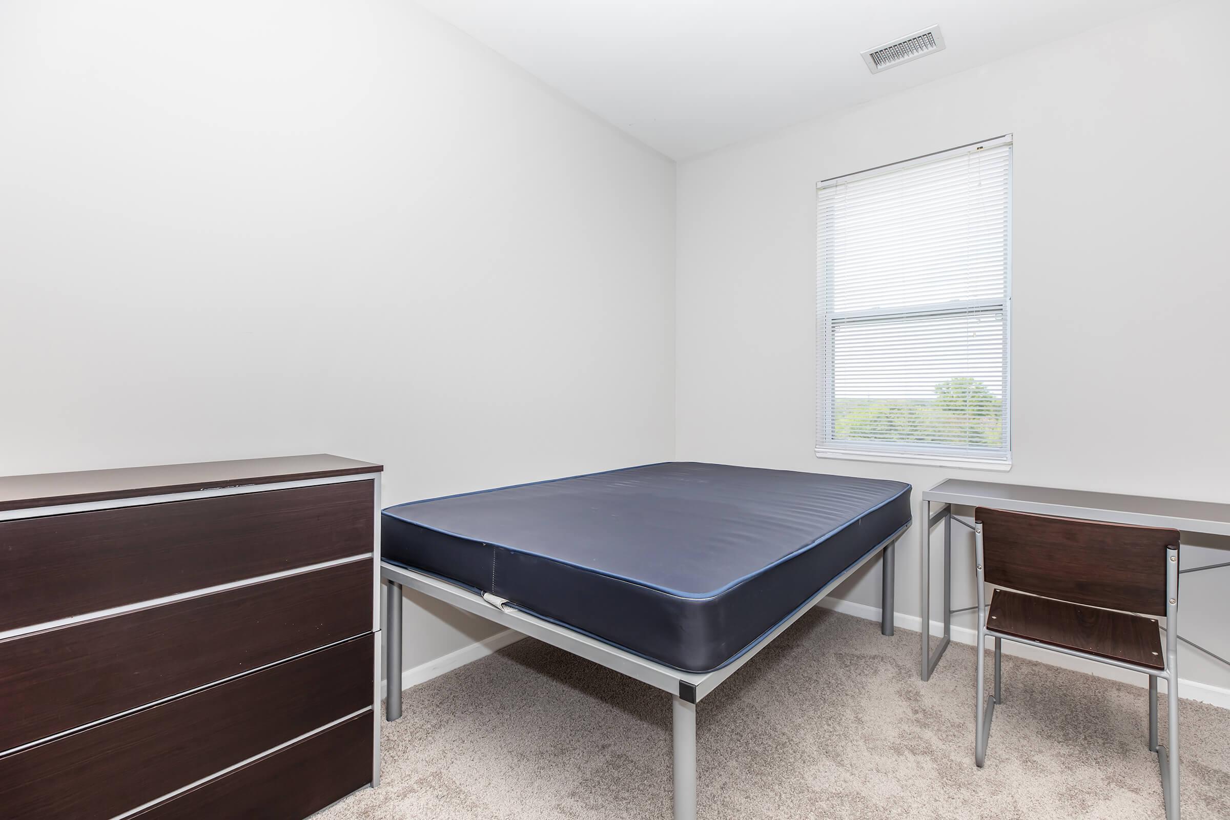 A simple, empty bedroom featuring a bed frame with a blue mattress, a dark wood dresser, and a small desk with a chair. The walls are plain, and there is a window with blinds allowing natural light to enter. The carpet is light-colored, giving a clean and spacious feel to the room.