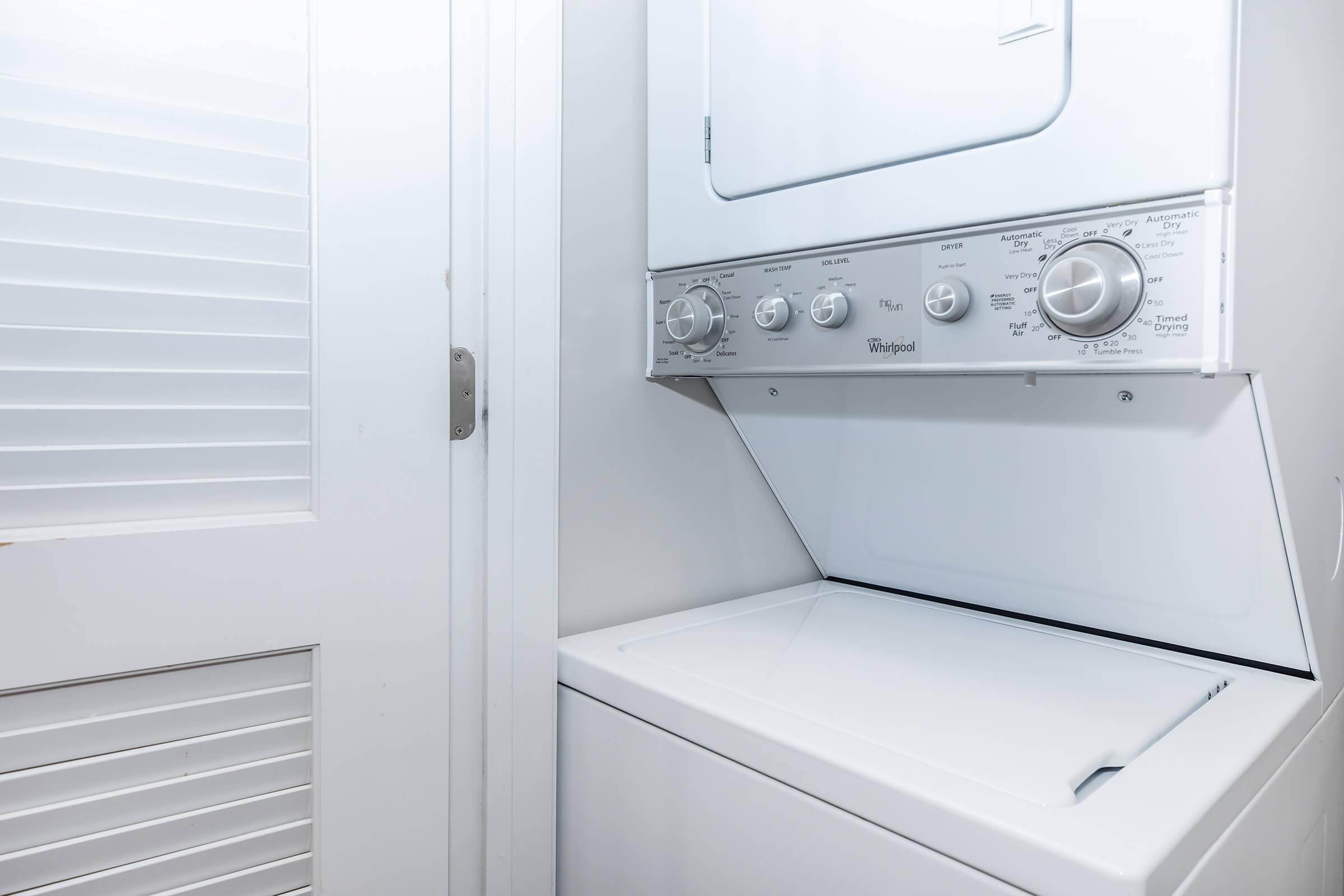 A stacked washer and dryer unit in a modern laundry area, featuring white appliances with dials and settings. The background includes a closed white door with vertical slats, paired with light-colored walls, creating a clean and organized appearance.