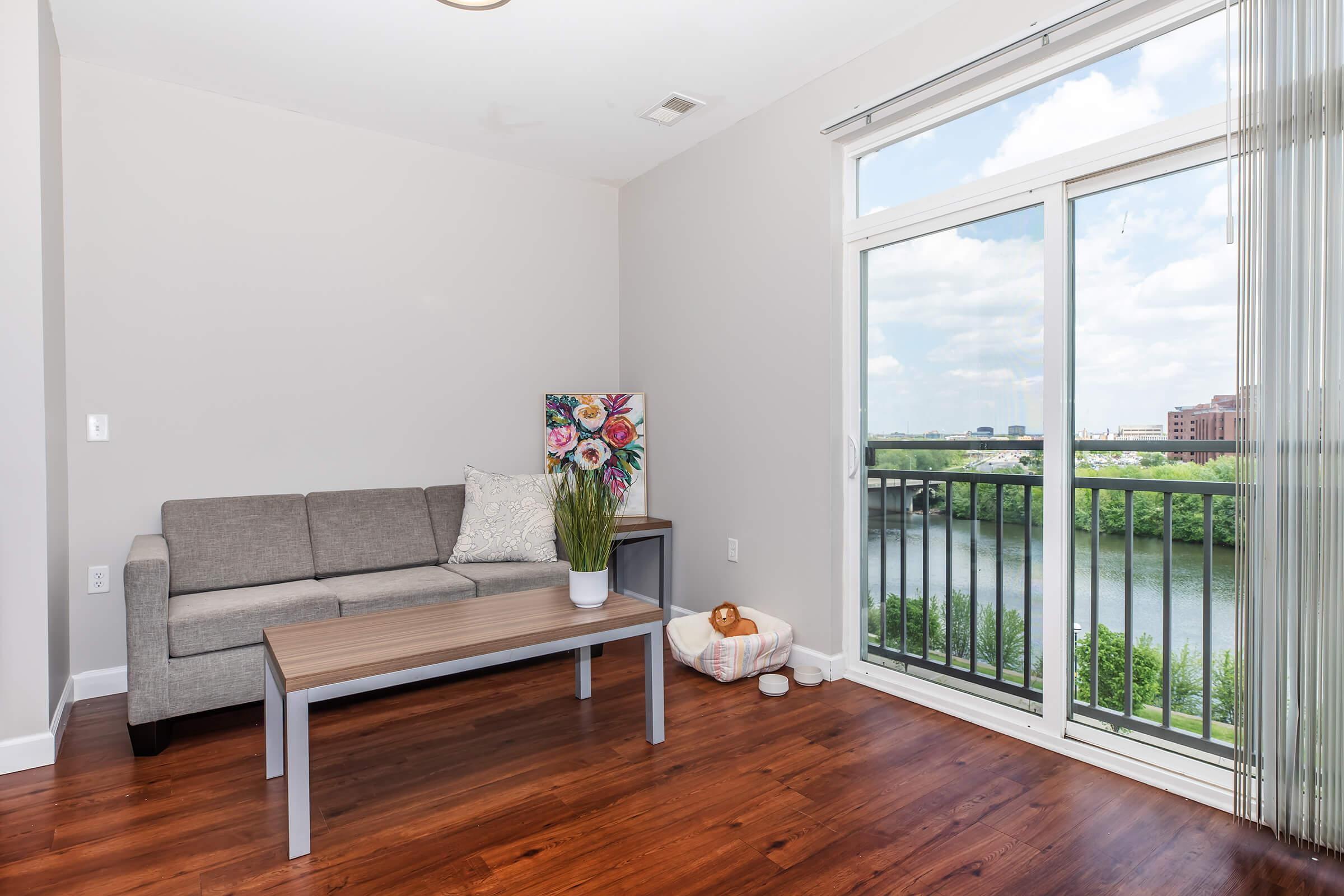 Modern living room featuring a gray sofa with decorative pillows, a wooden coffee table, and a potted plant. A large window with glass doors overlooks a river and greenery, letting in natural light. The floor is hardwood, creating a warm ambiance.