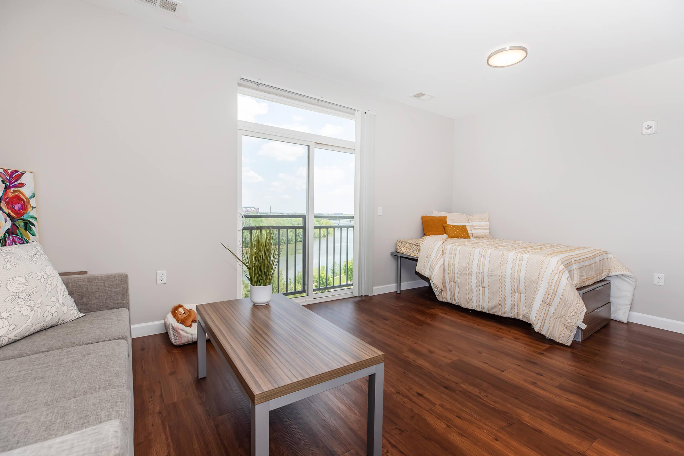 A modern, well-lit studio apartment featuring a grey sofa, a wooden coffee table, and a single bed with striped bedding. Large windows offer a view of greenery outside, allowing natural light to fill the space. The walls are painted in a soft color, creating a cozy atmosphere.