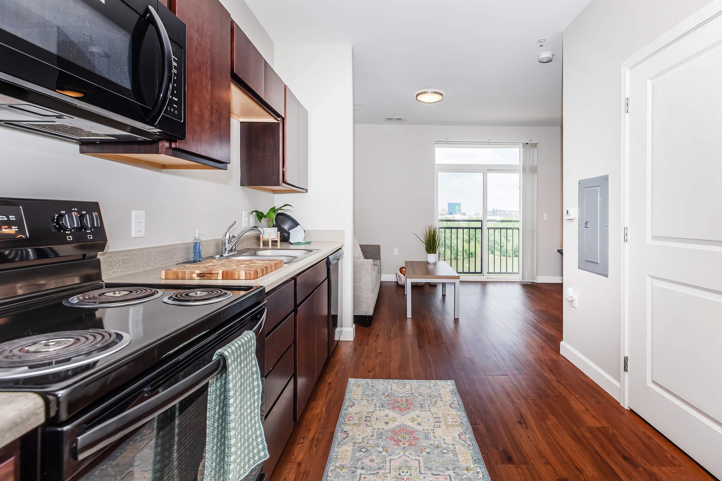 Modern kitchen with dark wood cabinetry, stainless steel appliances, and a countertop. In the background, a living room area features a small table and chair, with large windows showcasing outdoor greenery. Wood flooring adds warmth to the space.