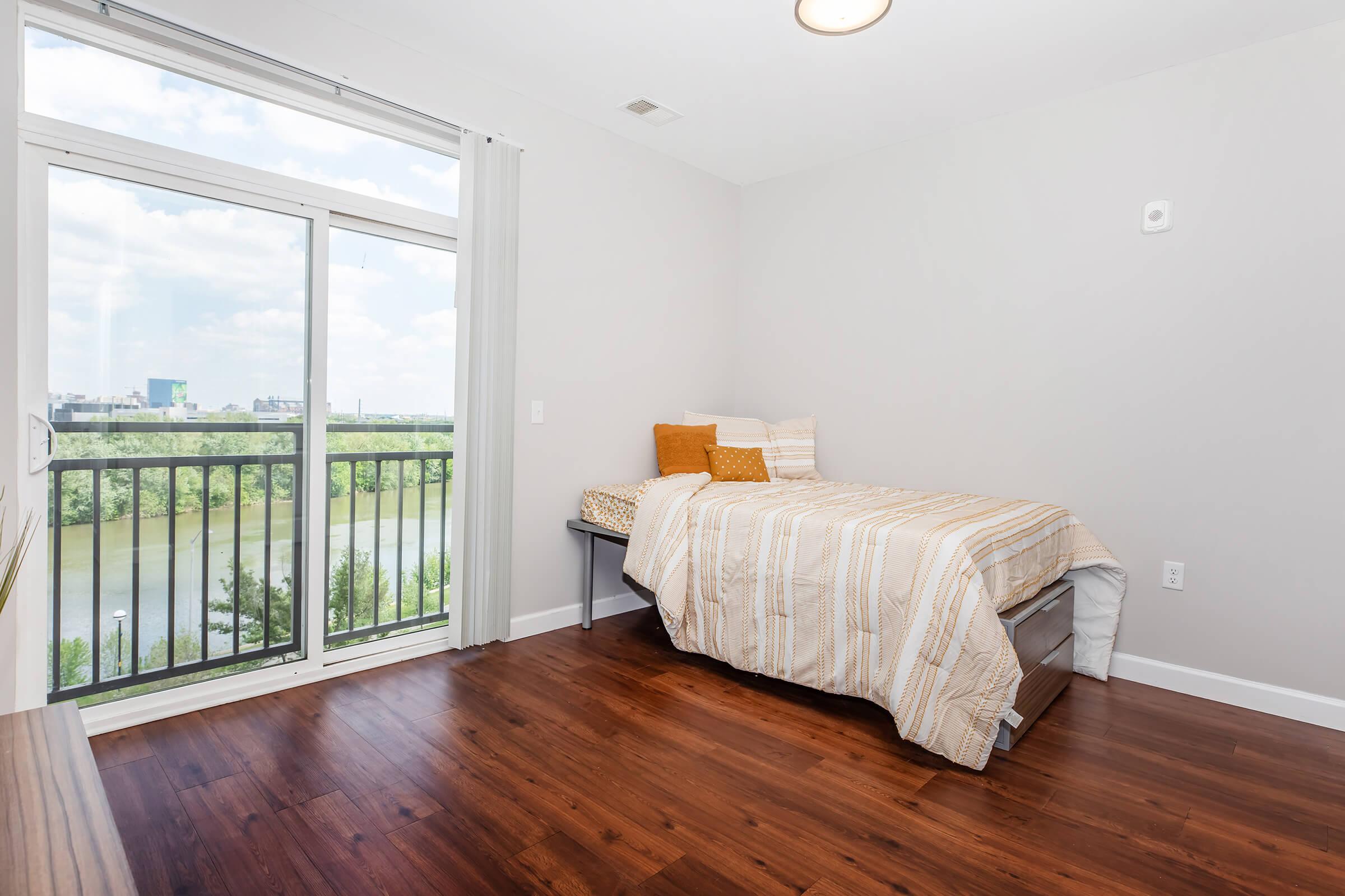 A cozy bedroom featuring a bed with striped bedding and orange pillows, a large window with a view of greenery and water, light gray walls, and wooden flooring. The room is well-lit with natural light and includes a small bedside table.