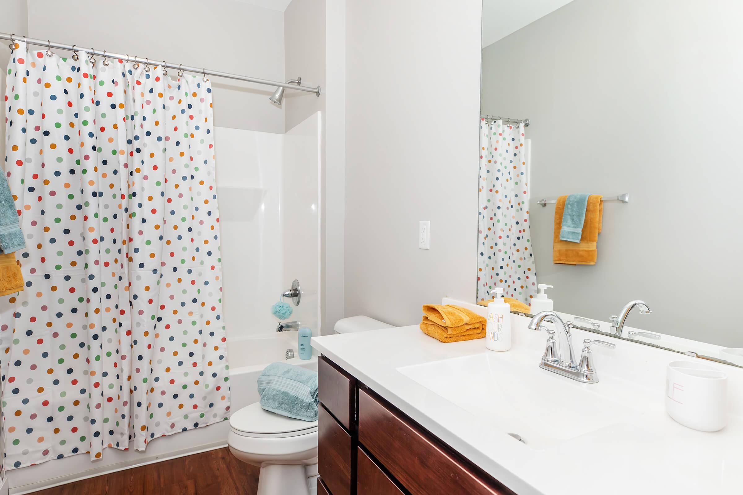 A bright and modern bathroom featuring a white tub with a polka-dot shower curtain, a clean vanity with a white countertop, and wooden cabinetry. Soft towels in shades of blue and yellow are neatly arranged on the counter and hanging on hooks, creating a welcoming and cheerful atmosphere.