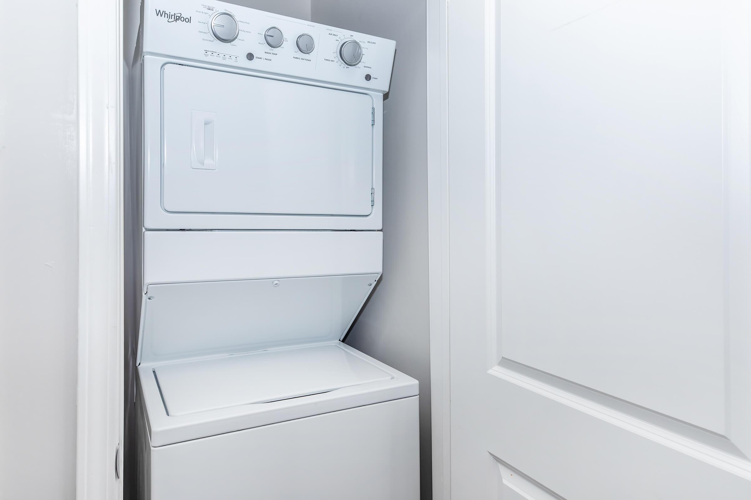 A stacked white washer and dryer unit installed in a narrow space, with a closed door in the background. The appliance features control knobs and a door on the front of the dryer. The surrounding walls are painted a light color, contributing to a clean, minimalist look.