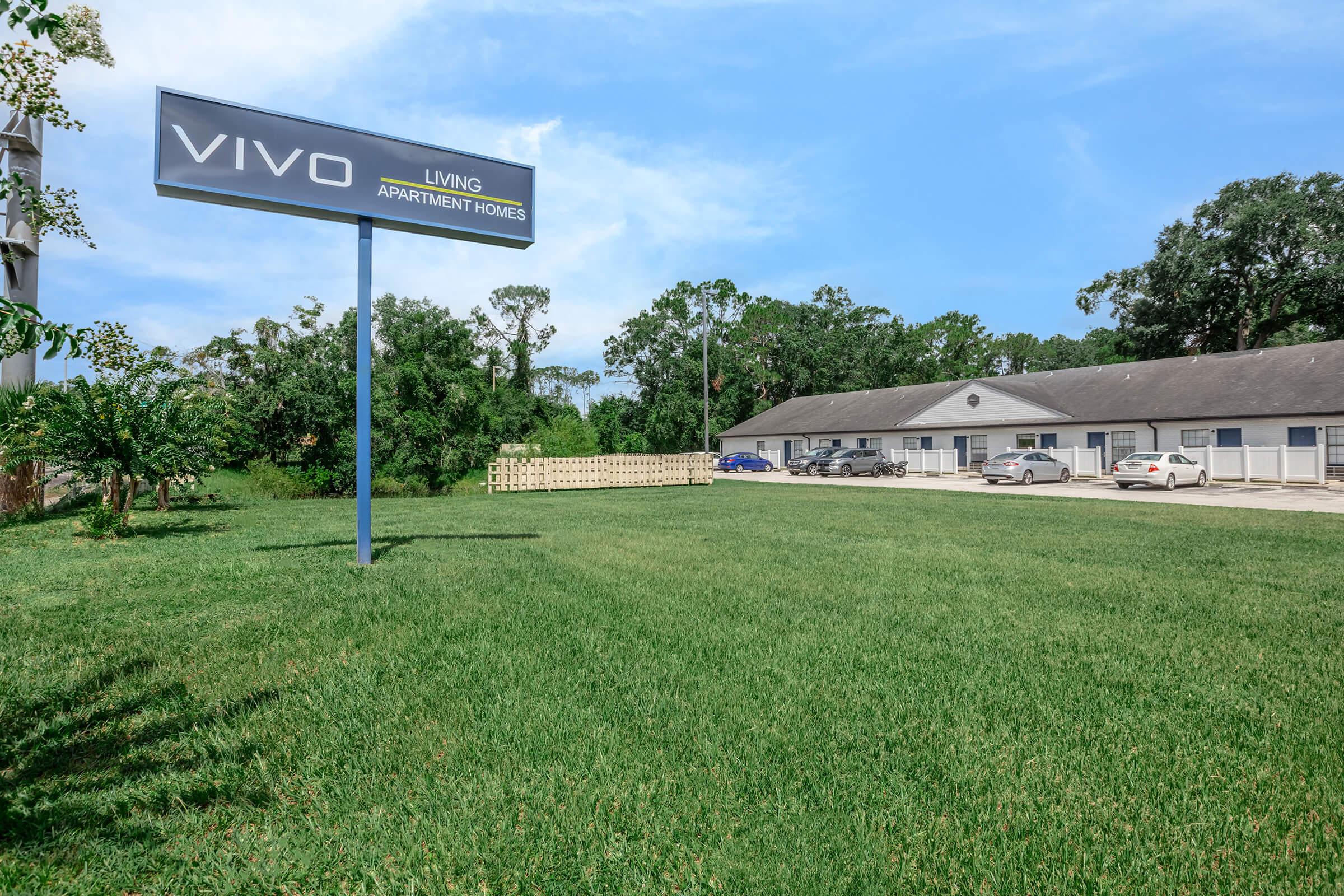 Sign for Vivo Living Apartment Homes in front of a grassy area, featuring a simple building with parking spaces and several cars. Lush green trees and blue sky in the background.