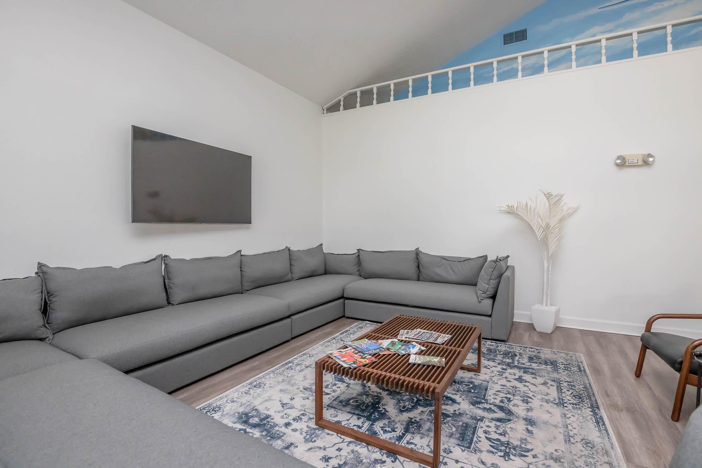 A modern living room featuring a large gray sectional sofa, a wooden coffee table with magazines on it, a wall-mounted TV, and a potted plant. The ceiling has a painted blue sky design, and there is a rug on the floor. The space is bright and inviting, with minimal decor.