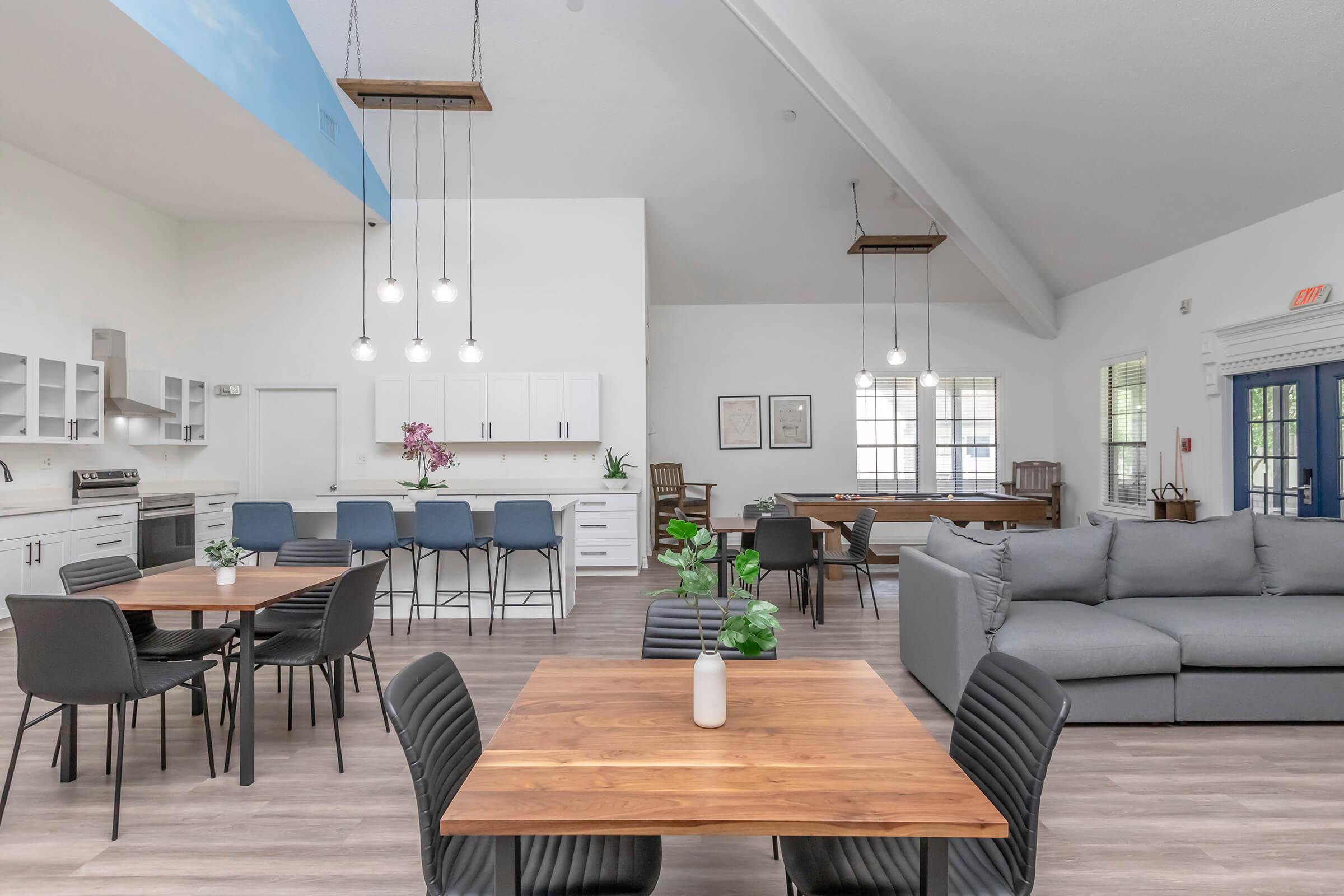 Brightly lit common area with modern furnishings. Features several wooden tables with black chairs, a large gray sectional sofa, and a kitchen with white cabinetry. Decorative elements include a vase of flowers and hanging pendant lights, creating a welcoming atmosphere. Large windows provide natural light.