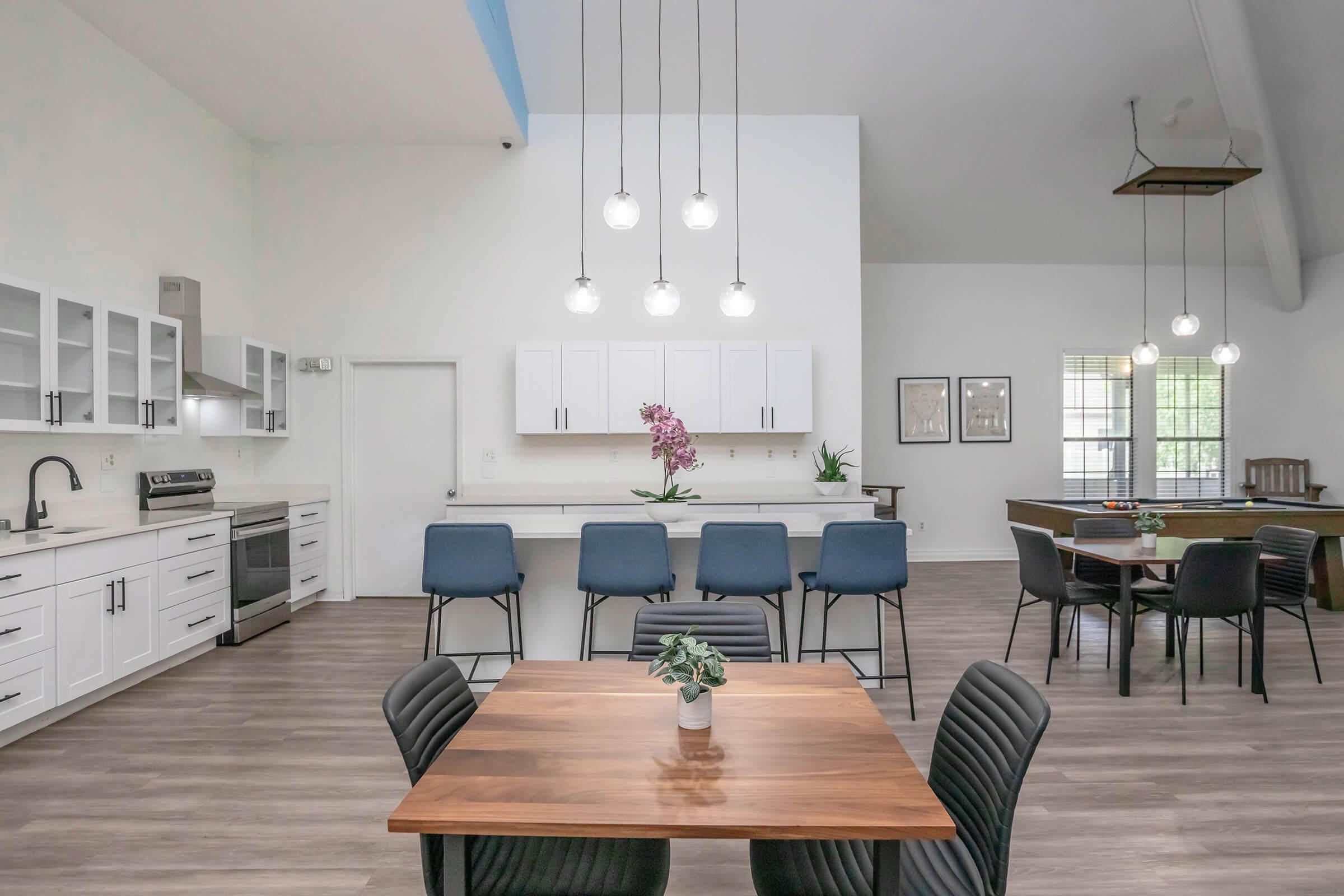 Bright and modern kitchen and dining area featuring a wooden table with black chairs, an island with bar stools, and sleek white cabinetry. Large windows allow natural light to fill the space, complemented by stylish pendant lighting. A touch of greenery adds warmth to the contemporary design.