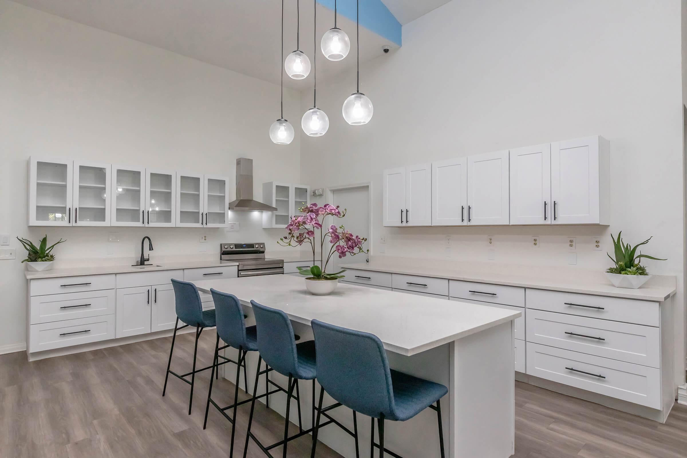Modern kitchen featuring white cabinetry, a large island with seating for four, and pendant lights hanging from the ceiling. Glass-front cabinets and a stainless steel stove add to the sleek design. Decorated with green plants and a vase of pink orchids on the island, creating a bright and inviting atmosphere.