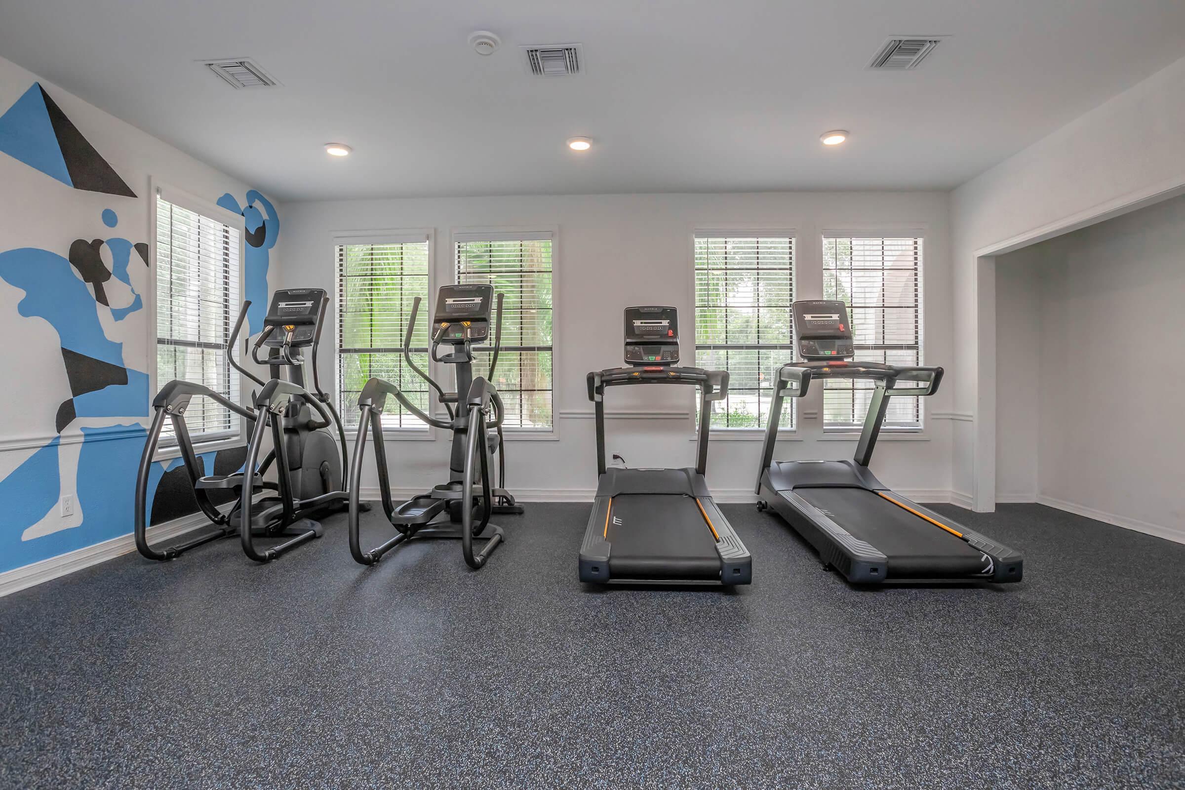 A fitness room featuring two treadmills and two elliptical machines, with large windows allowing natural light. There is a colorful wall mural depicting a person exercising, enhancing the energetic atmosphere. The floor is covered in dark rubber flooring for safety and comfort.