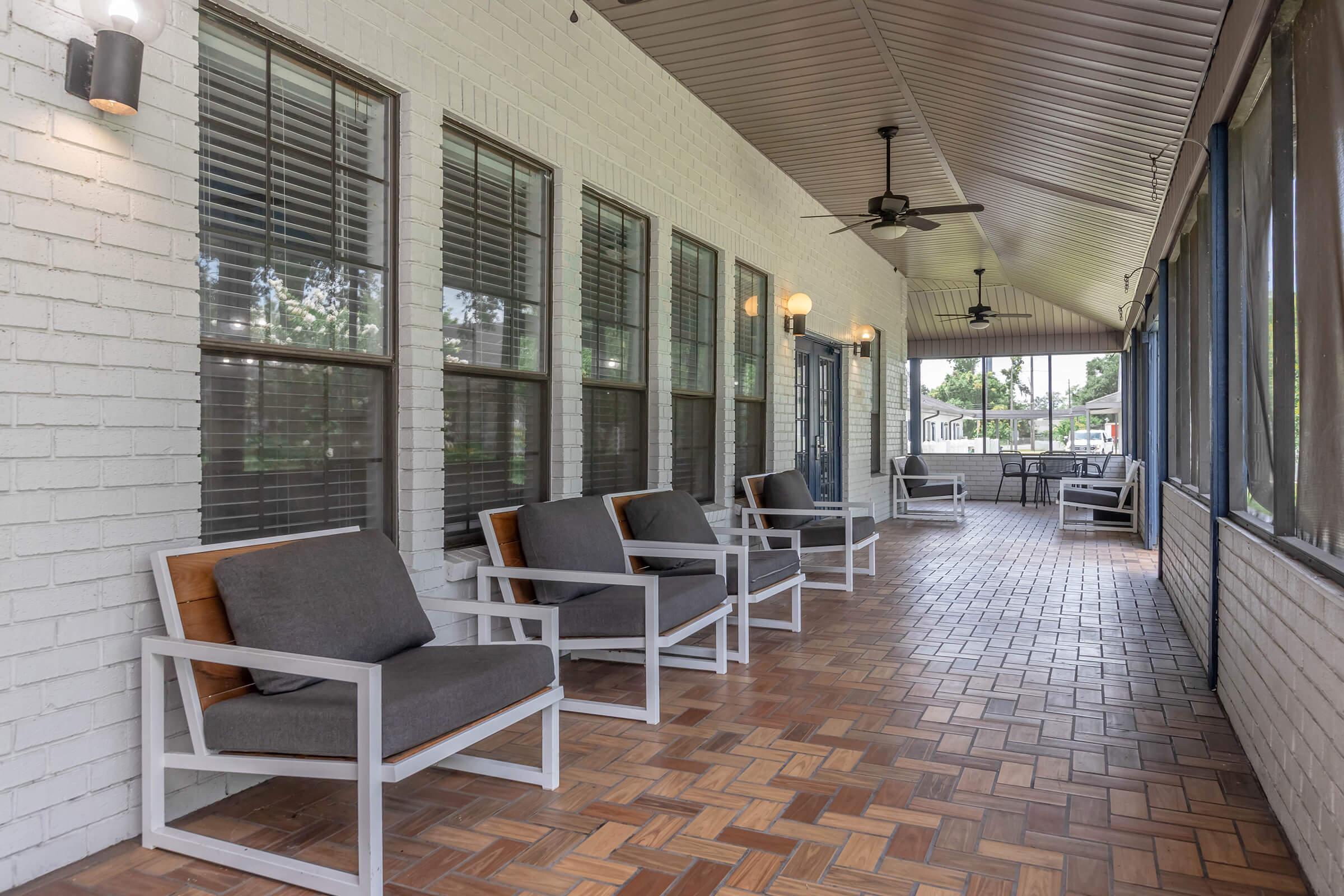 A spacious porch with a tiled floor and large windows. There are several modern gray chairs with wooden accents arranged along the sides. Ceiling fans are installed, and the environment appears bright and inviting, with greenery visible outside.