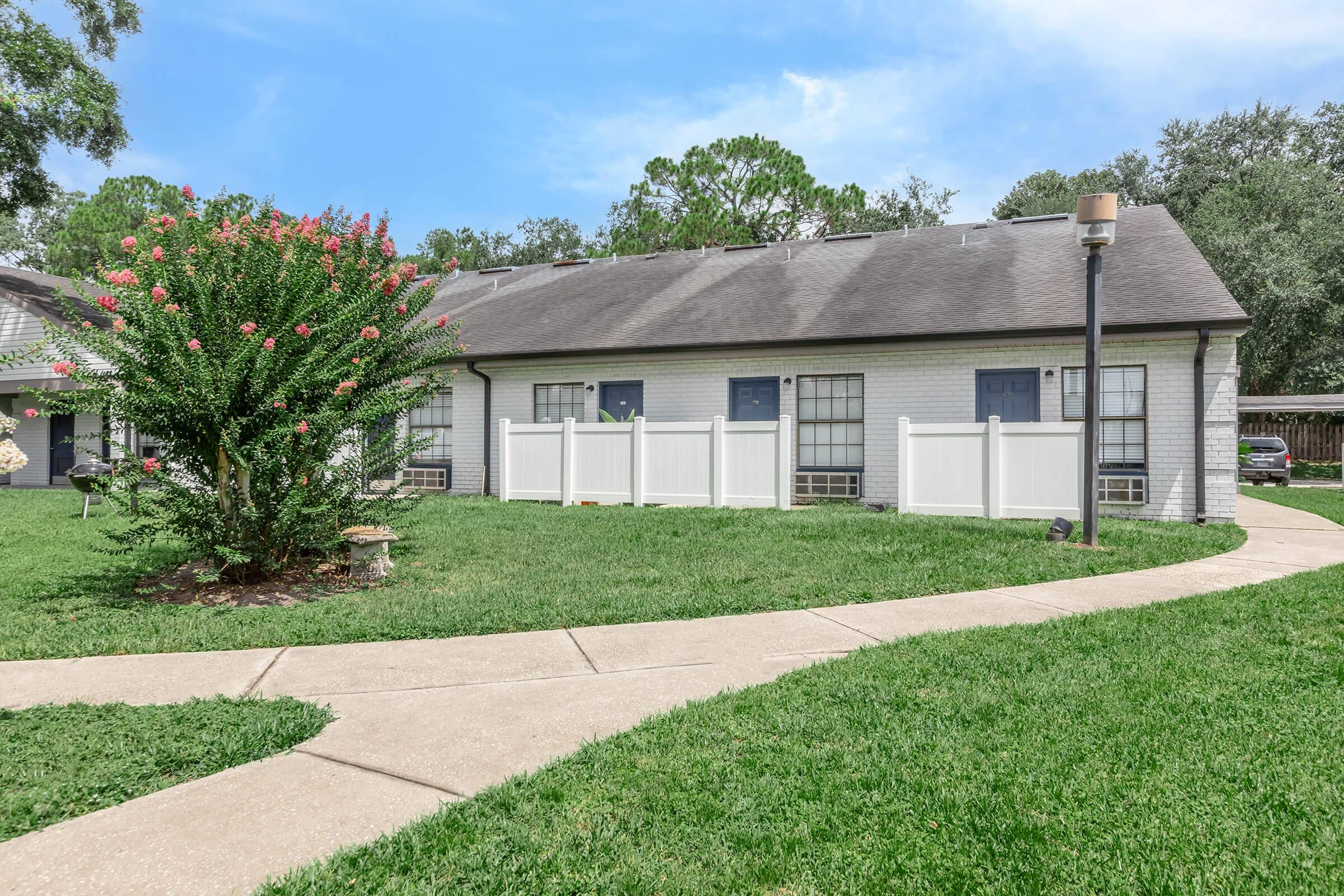 A suburban home with a white picket fence surrounded by green grass. The pathway leads to the front door, and there are trees and shrubs in the front yard under a clear blue sky. The house has a simple design with multiple windows.