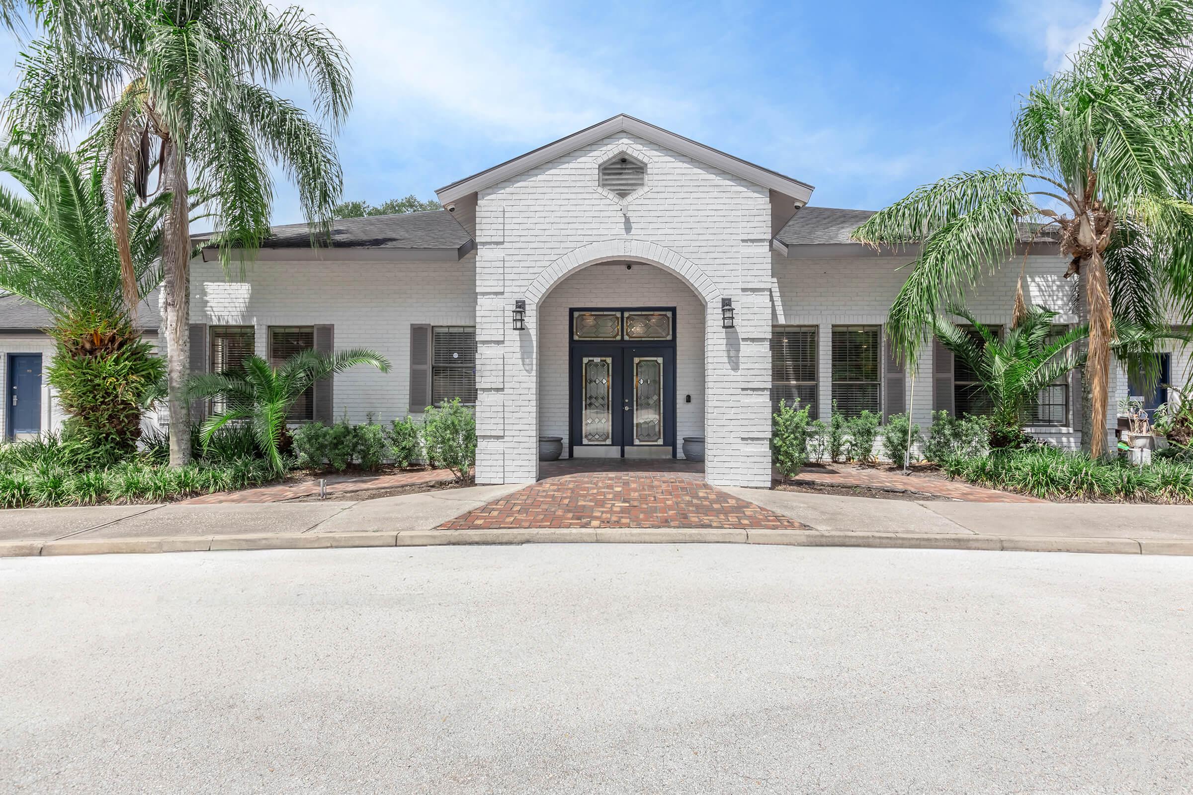 A modern single-story building with a white exterior and arched entrance. The entrance features large glass doors and is flanked by palm trees and lush greenery. A brick pathway leads to the entrance, while a wide, clear road runs in front of the building under a blue sky.
