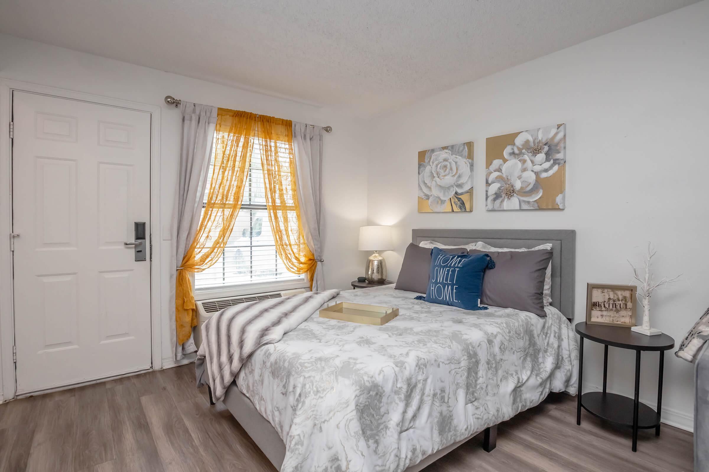 A cozy bedroom featuring a bed with gray and white bedding, decorative pillows, and a tray on top. There is a nightstand beside the bed, a lamp, and floral wall art. Natural light streams in through a window adorned with sheer curtains and vibrant yellow drapes. A door leads to the entrance of the room.