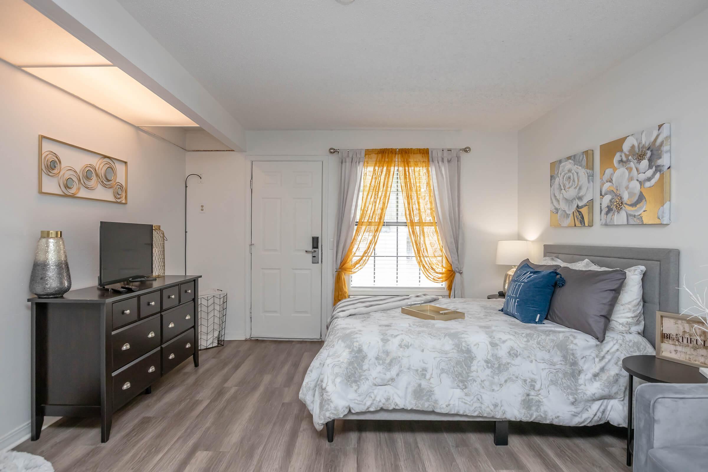 A cozy bedroom featuring a bed with decorative pillows, a nightstand, and a flat-screen TV on a dresser. Natural light filters through a window adorned with yellow curtains. The room has a neutral color palette with floral wall art, creating a welcoming atmosphere.